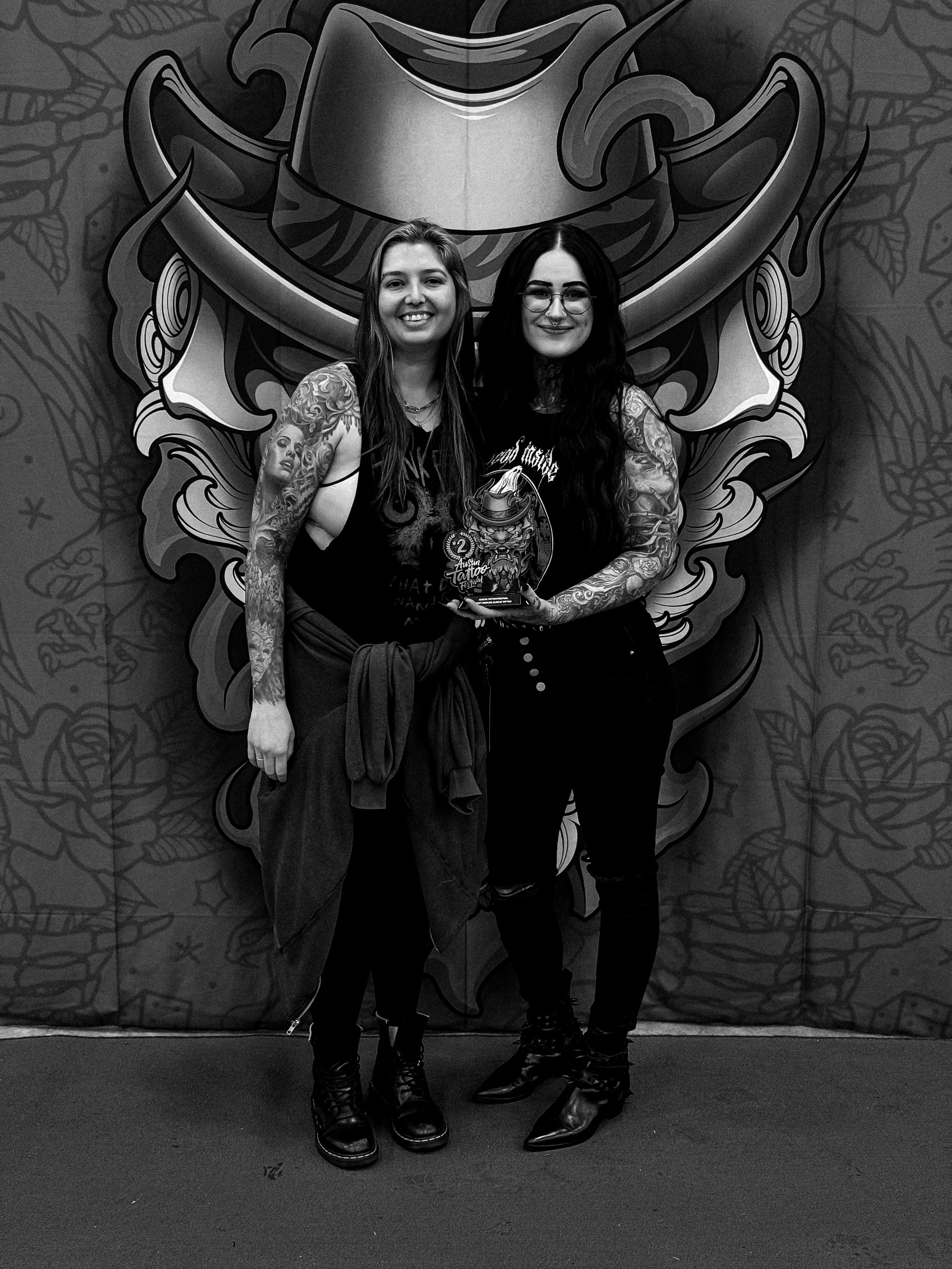 a photo of two tattooed women, one being a female tattoo artist holding a tattoo award she won for a tattoo on the other woman's arm. Taken at the Austin Tattoo Show.