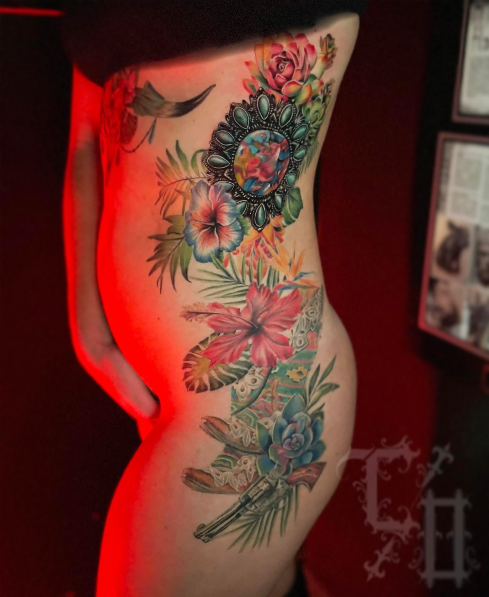Close-up of a person's side torso featuring a colorful tattoo of flowers, leaves, butterflies, and a vintage-style mirror.