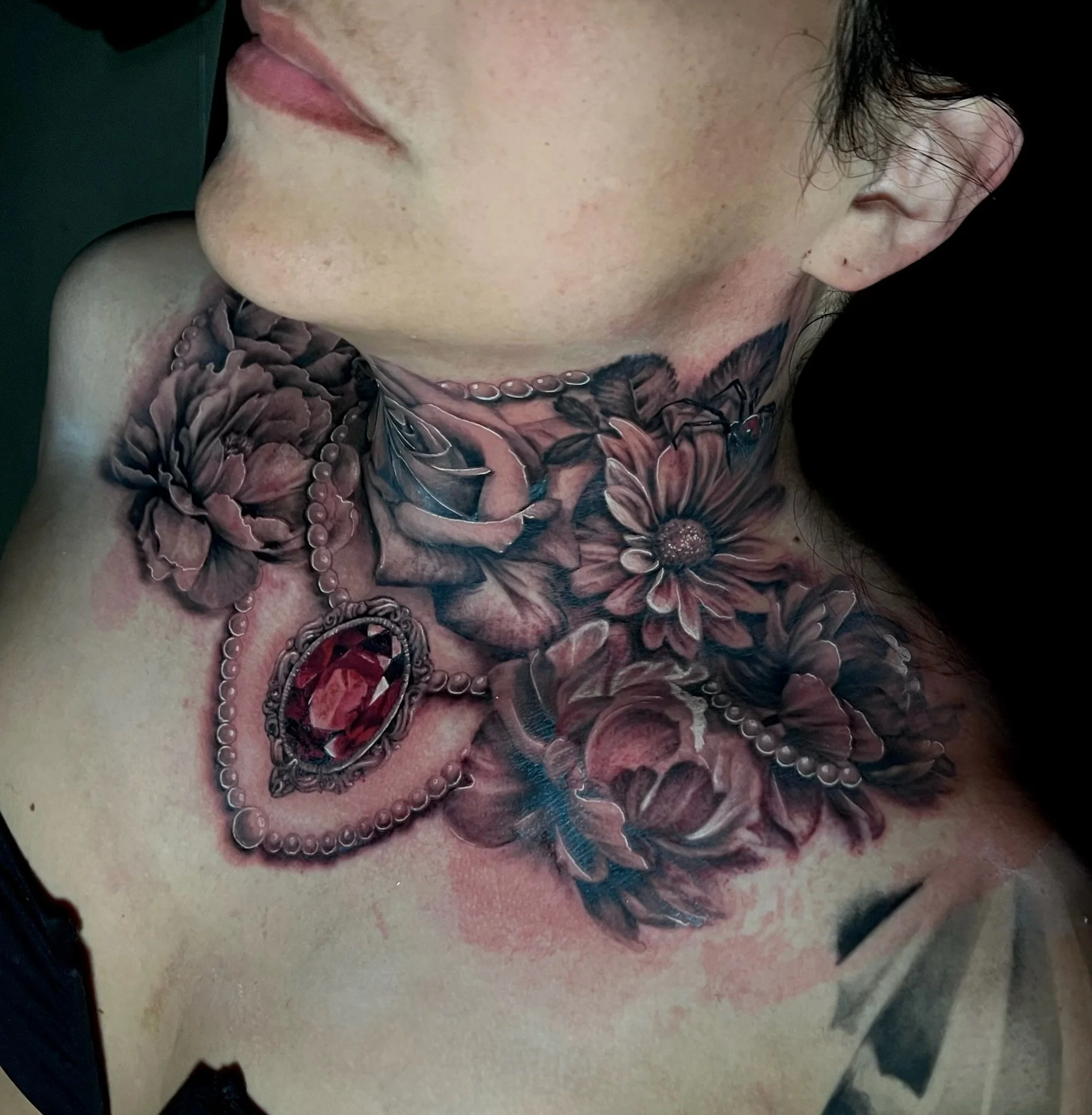 Close-up of a person's chest and neck tattoos featuring flowers, leaves, pearls, and a large red gem pendant.