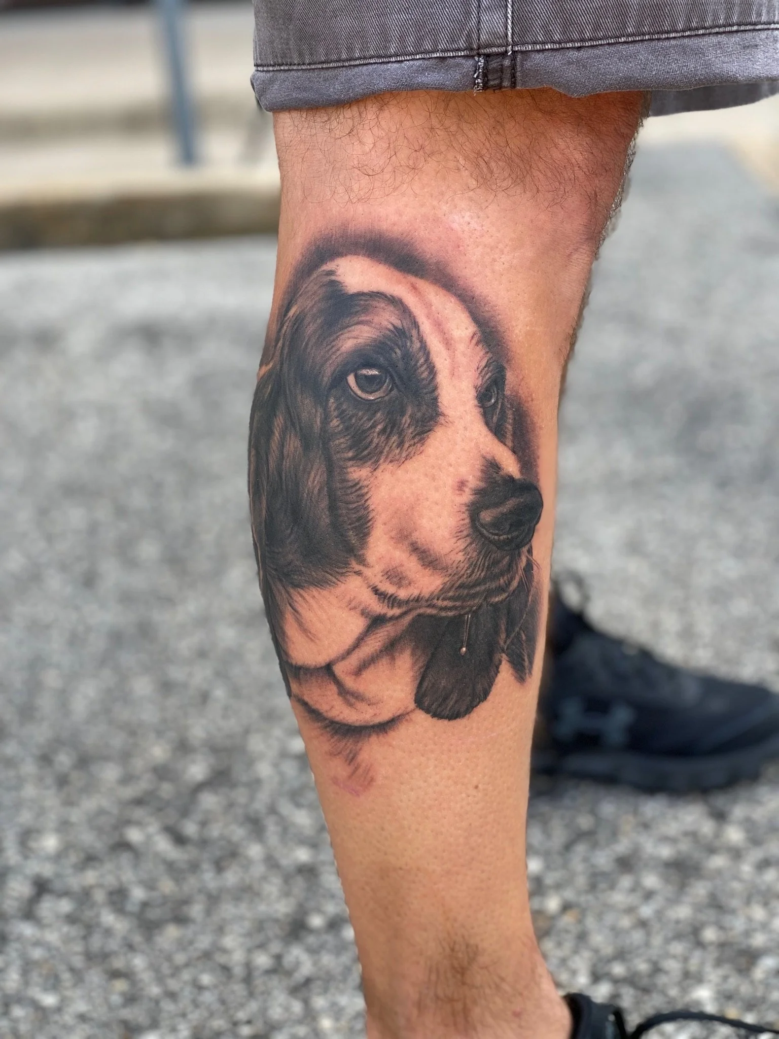 Tattoo of a dog's face on a person's calf. The dog appears to be a young, floppy-eared breed, possibly a retriever or similar type, in black and gray ink.