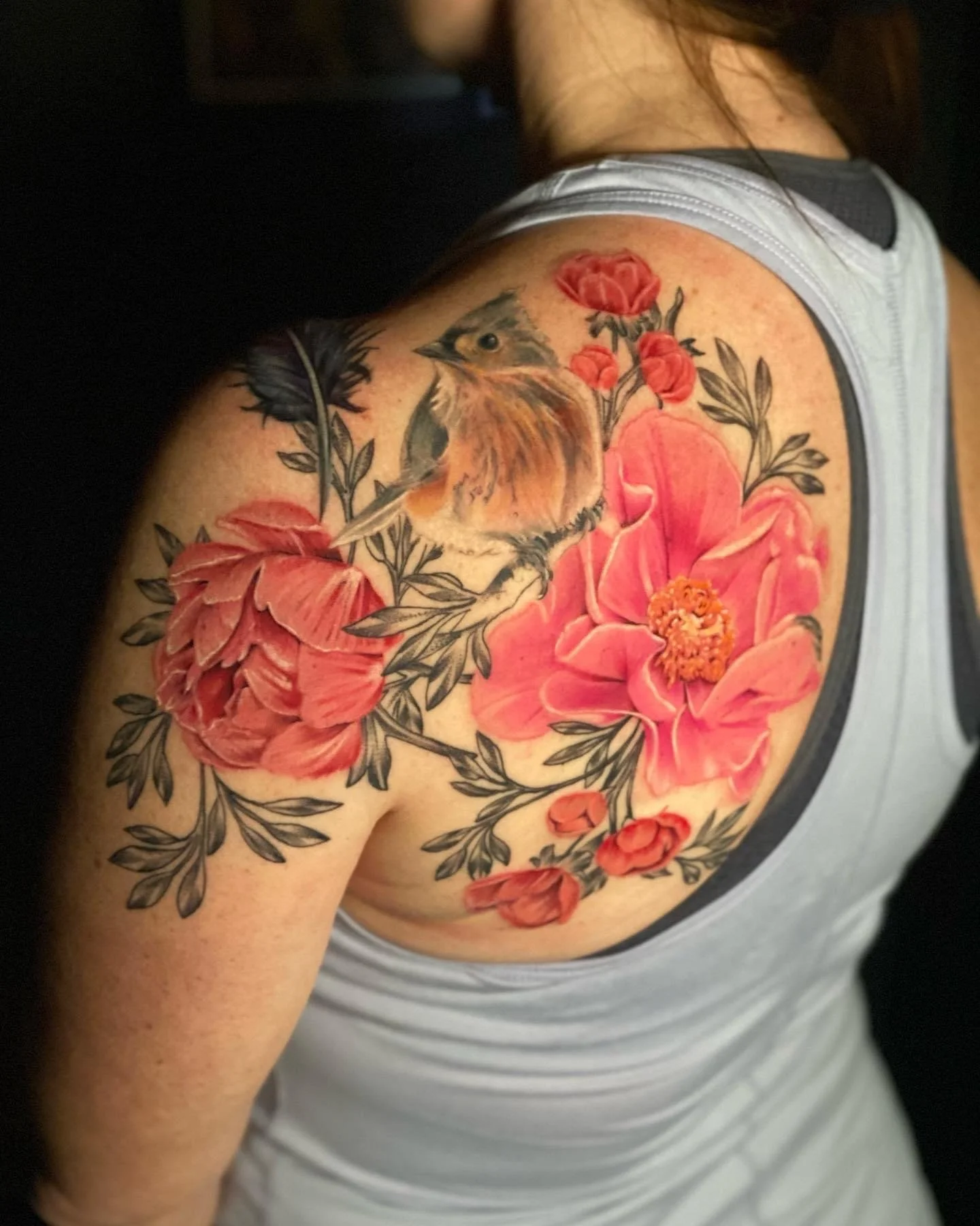 Colorful tattoo of a bird perched on a branch with pink flowers on a person's shoulder and upper chest.