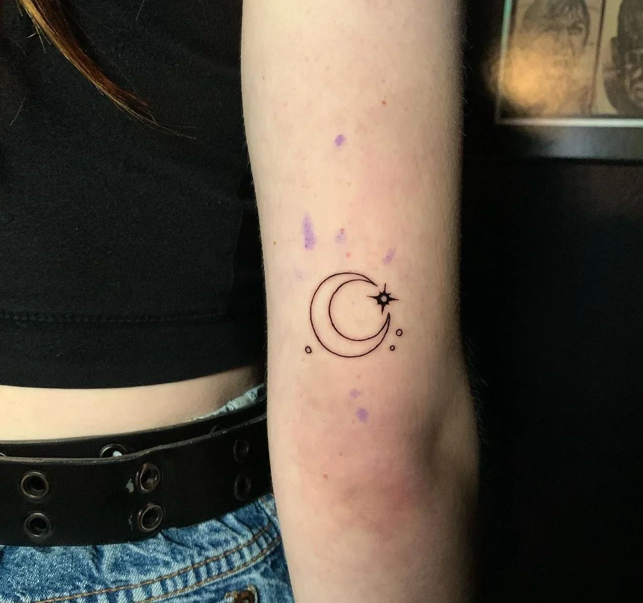 Tattoo of a crescent moon with a star and small circles on a person's forearm.