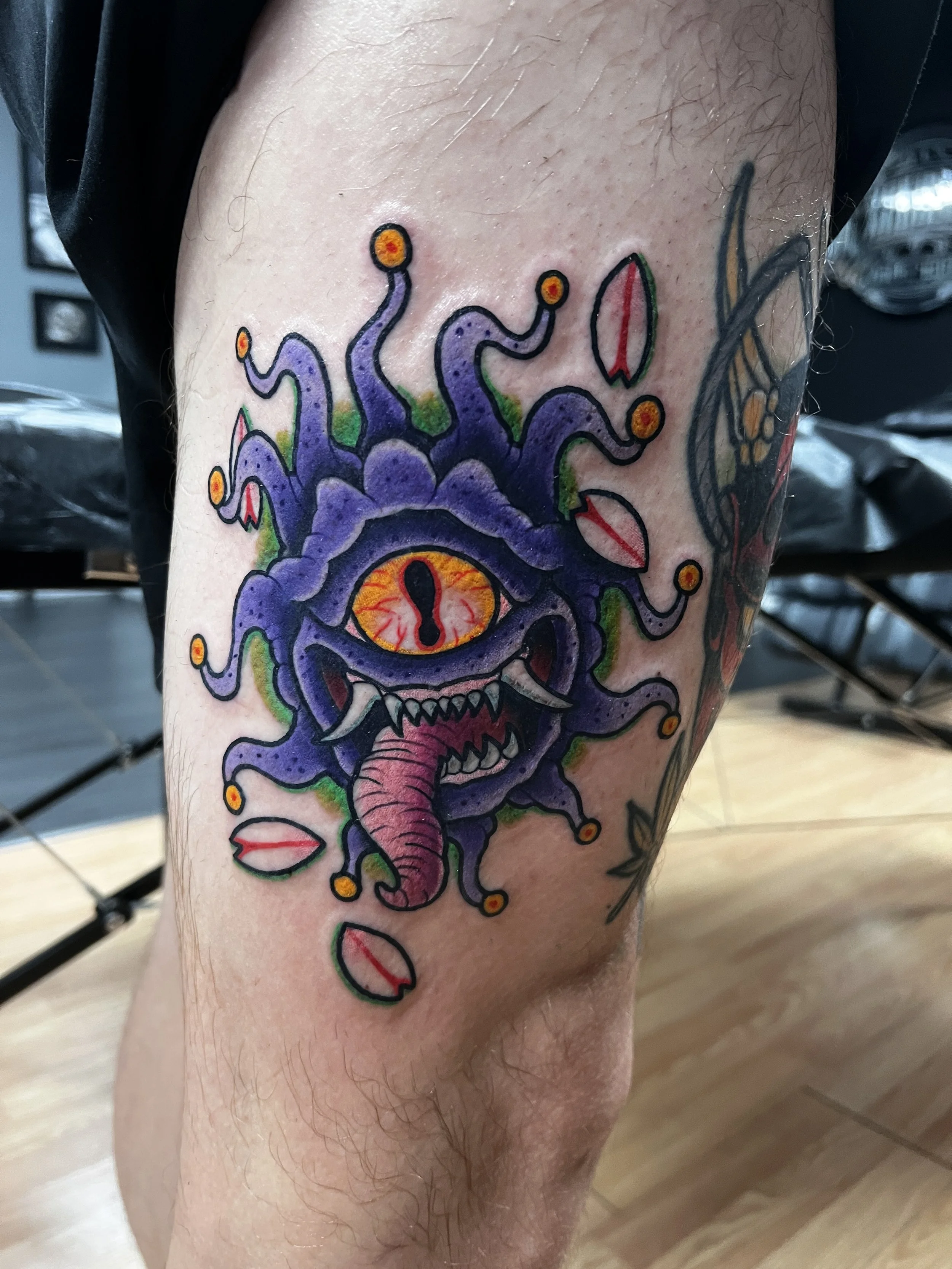 Colorful tattoo of a stylized, menacing creature with an eye, tentacle-like appendages, and sharp teeth, on a person's leg.