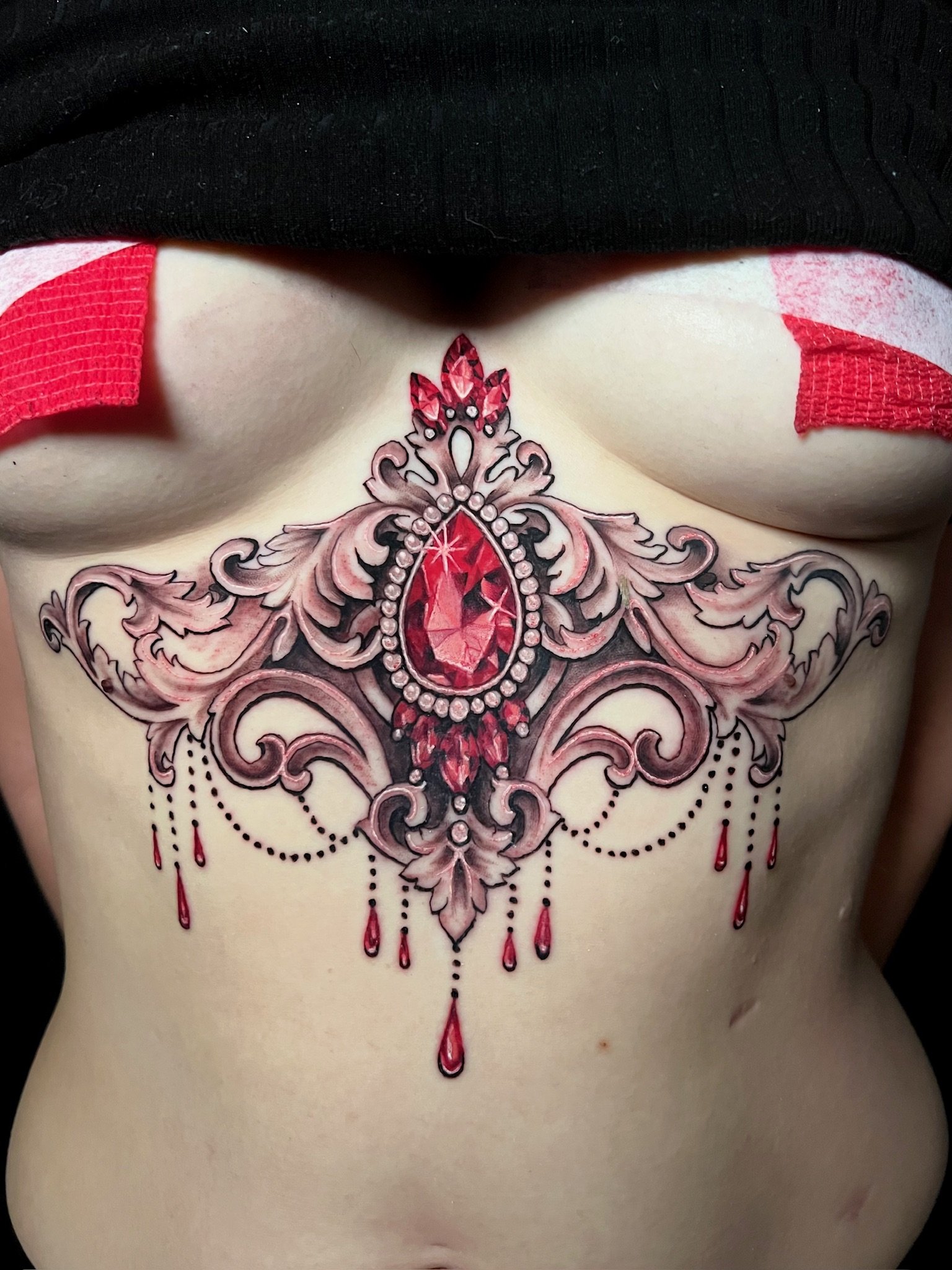 Colorful chest tattoo of a large red gemstone surrounded by ornate gray and black designs with red accents and hanging red teardrop shapes.