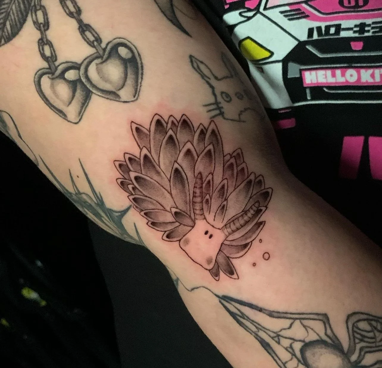 Tattoo on a person's arm featuring a cartoonish mushroom with a face, surrounded by other tattoos including hearts with chains and a Hello Kitty design on clothing nearby.