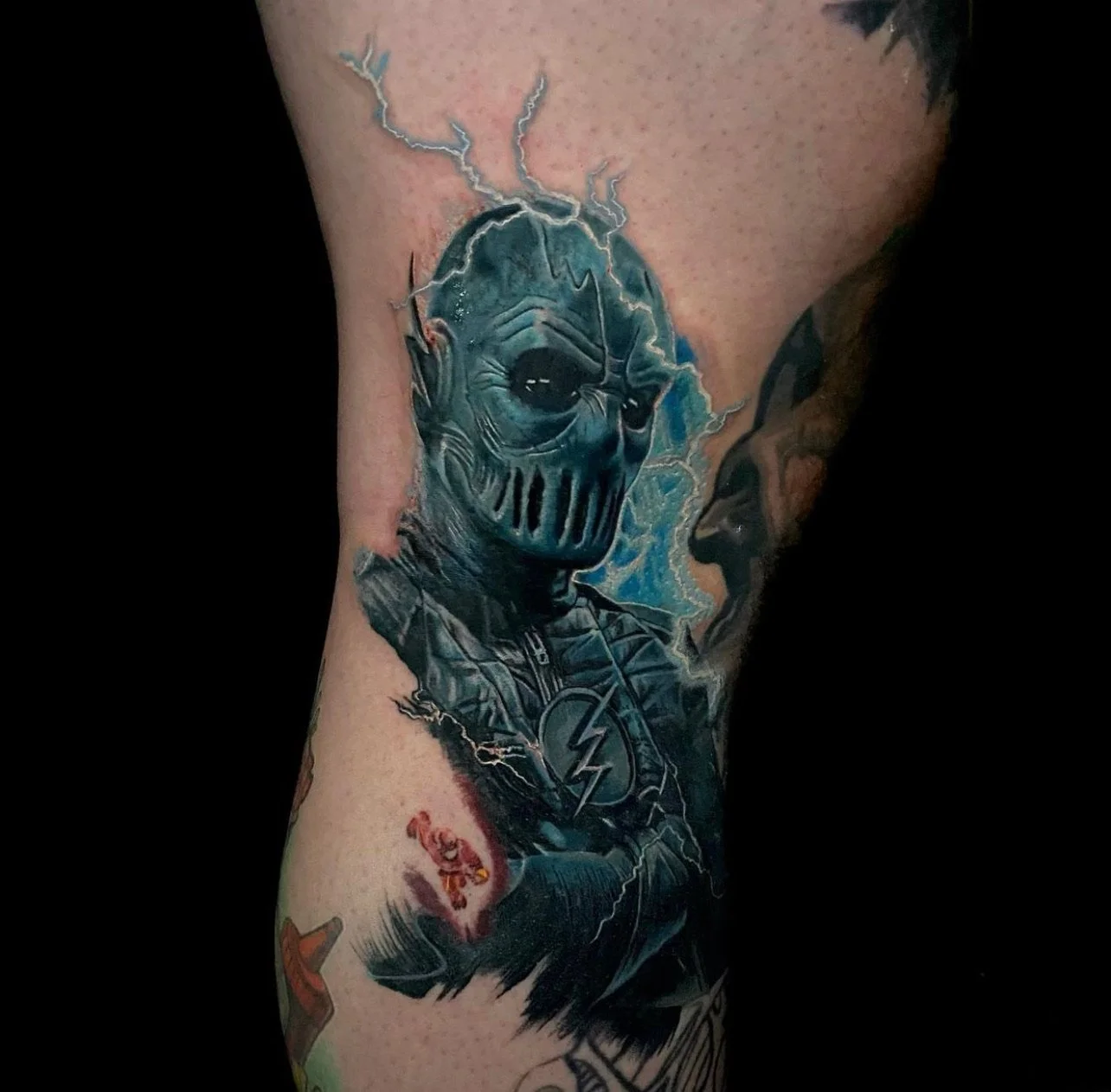 Tattoo of a humanoid figure with a metallic skull mask, lightning and electric effects around the head, wearing a superhero jacket with a lightning bolt symbol, and a small red creature below.
