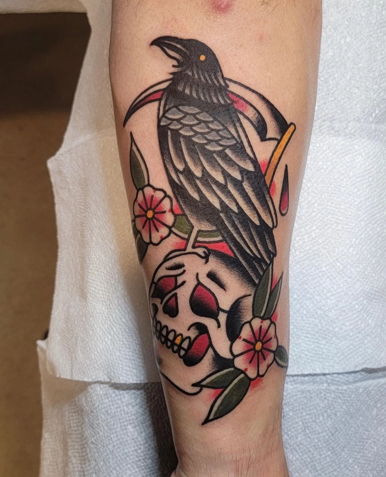A tattoo featuring a black bird, a skull, and pink flowers with green leaves.