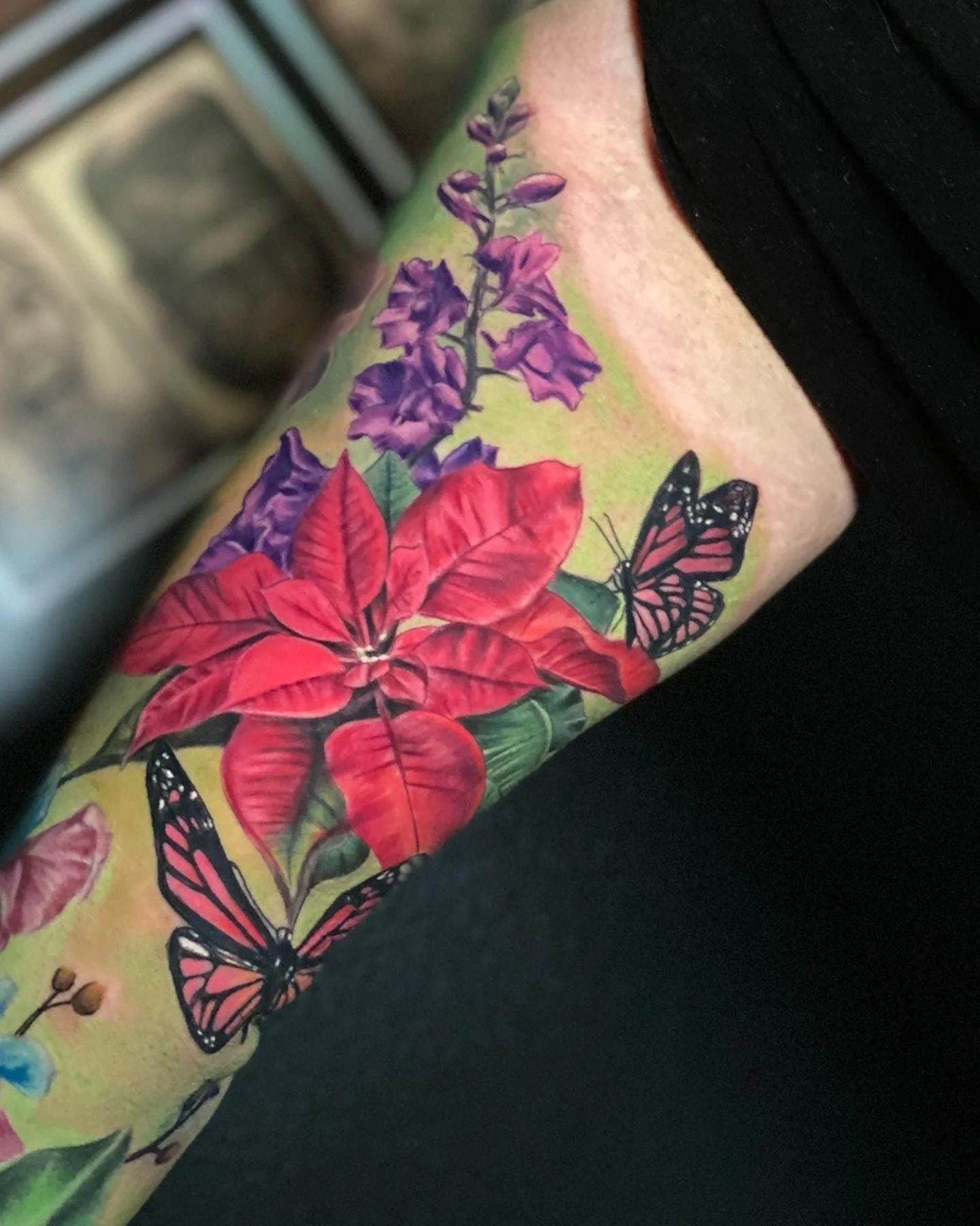 Colorful tattoo of purple, red, and green flowers with two black, pink, and white butterflies on a person's arm.