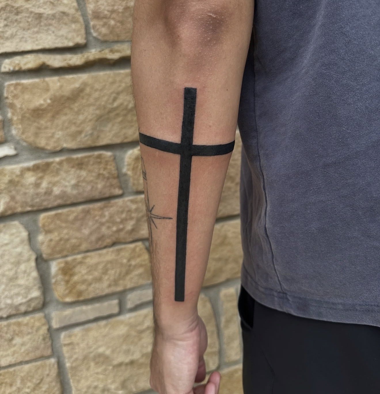 A person's arm with a black ink tattoo of a vertical and horizontal line forming a cross, with a brick wall in the background.