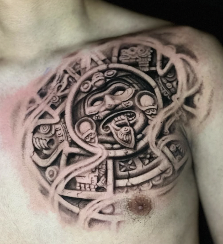 A black and grey tattoo on a person's chest depicting a Mayan or Aztec style design with a central face, skulls, and intricate patterns.