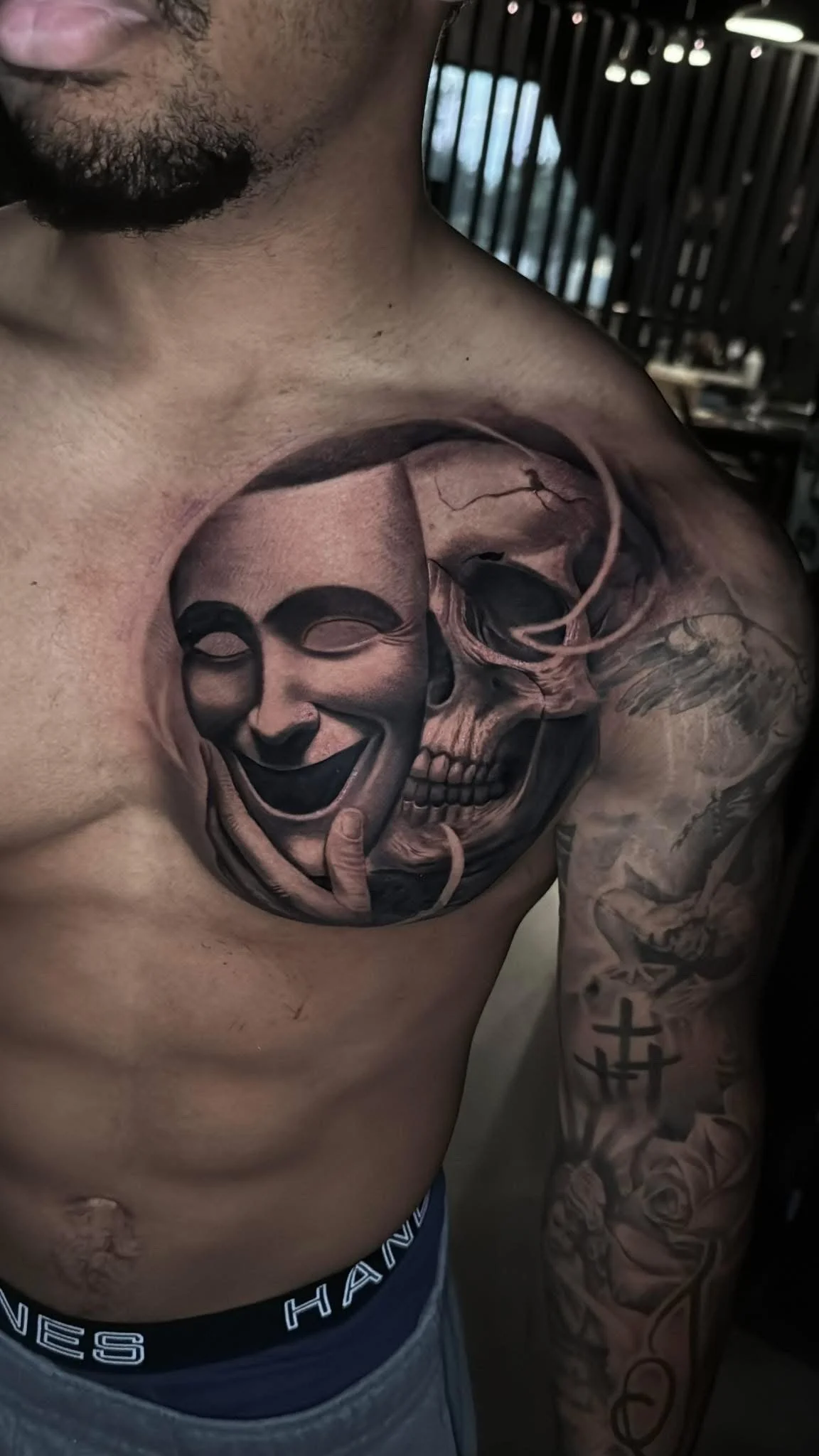 Tattoo of a theatrical comedy mask and a skull on a man's chest. The mask is smiling, and the skull appears realistic, blending into the mask.