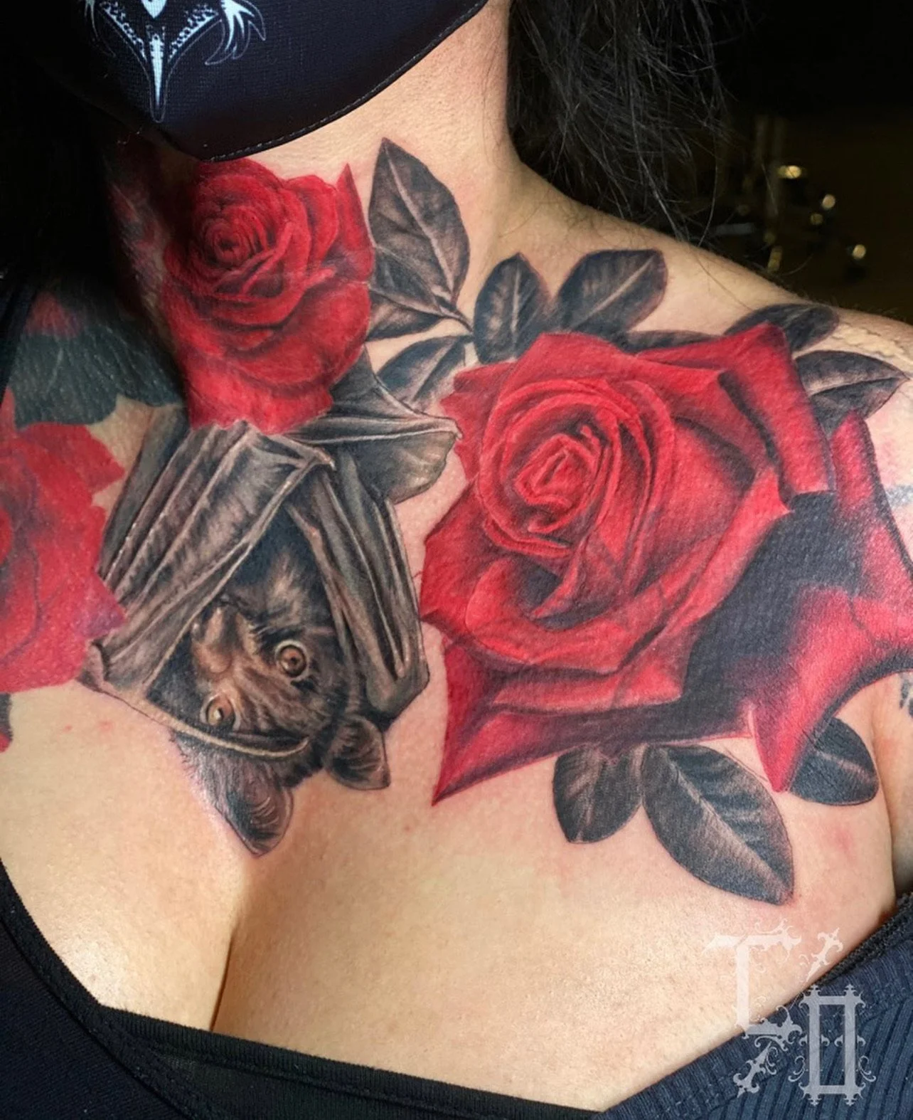Tattoo of red roses and black leaves on a person's shoulder and chest, with a bat hanging upside down between the roses.