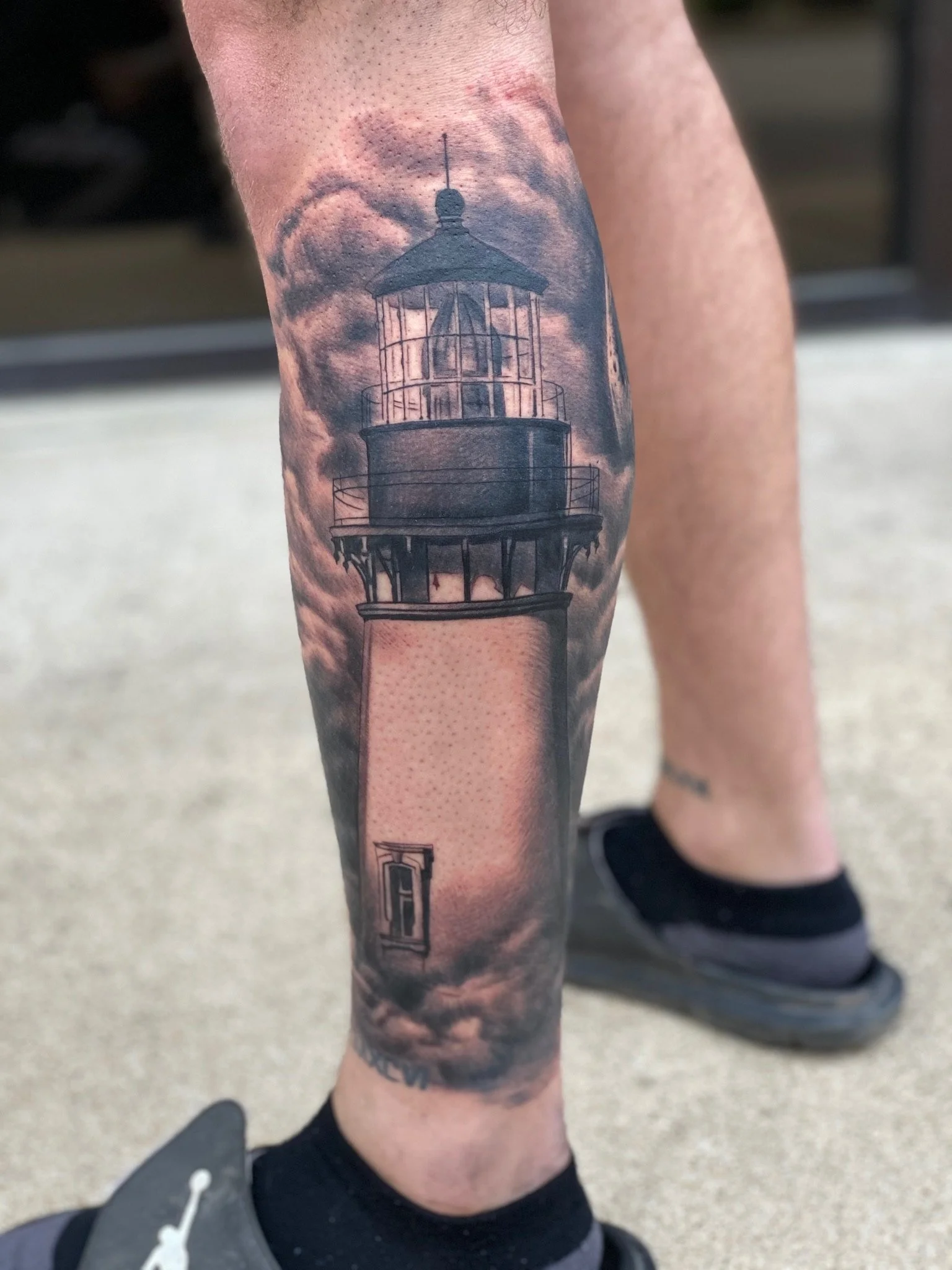 Black and gray tattoo of a lighthouse on leg with cloudy sky background.