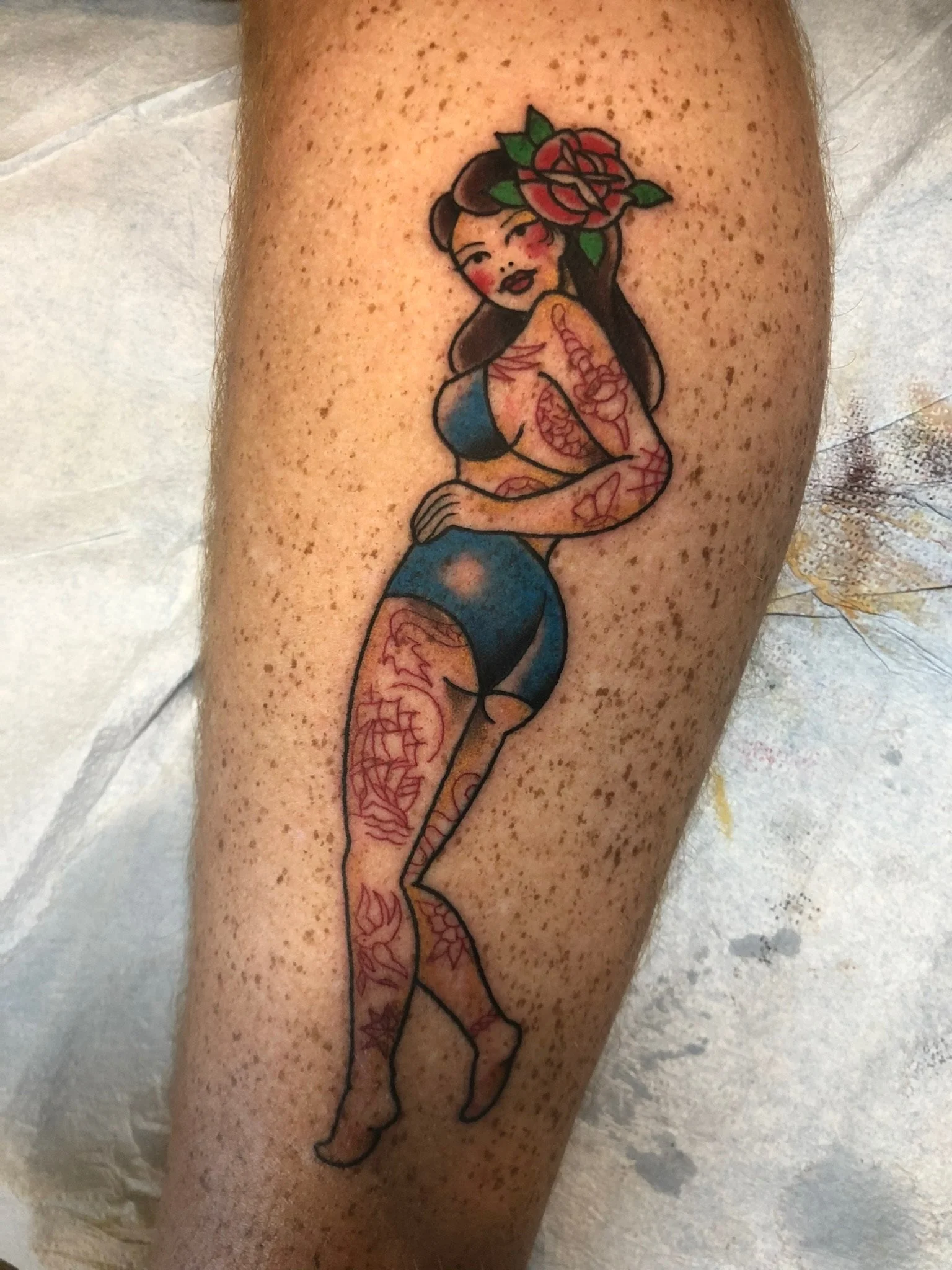 Tattoo of a pin-up style woman with a rose in her hair, wearing a blue top and shorts, with floral tattoos on her arms, legs, and shoulders.