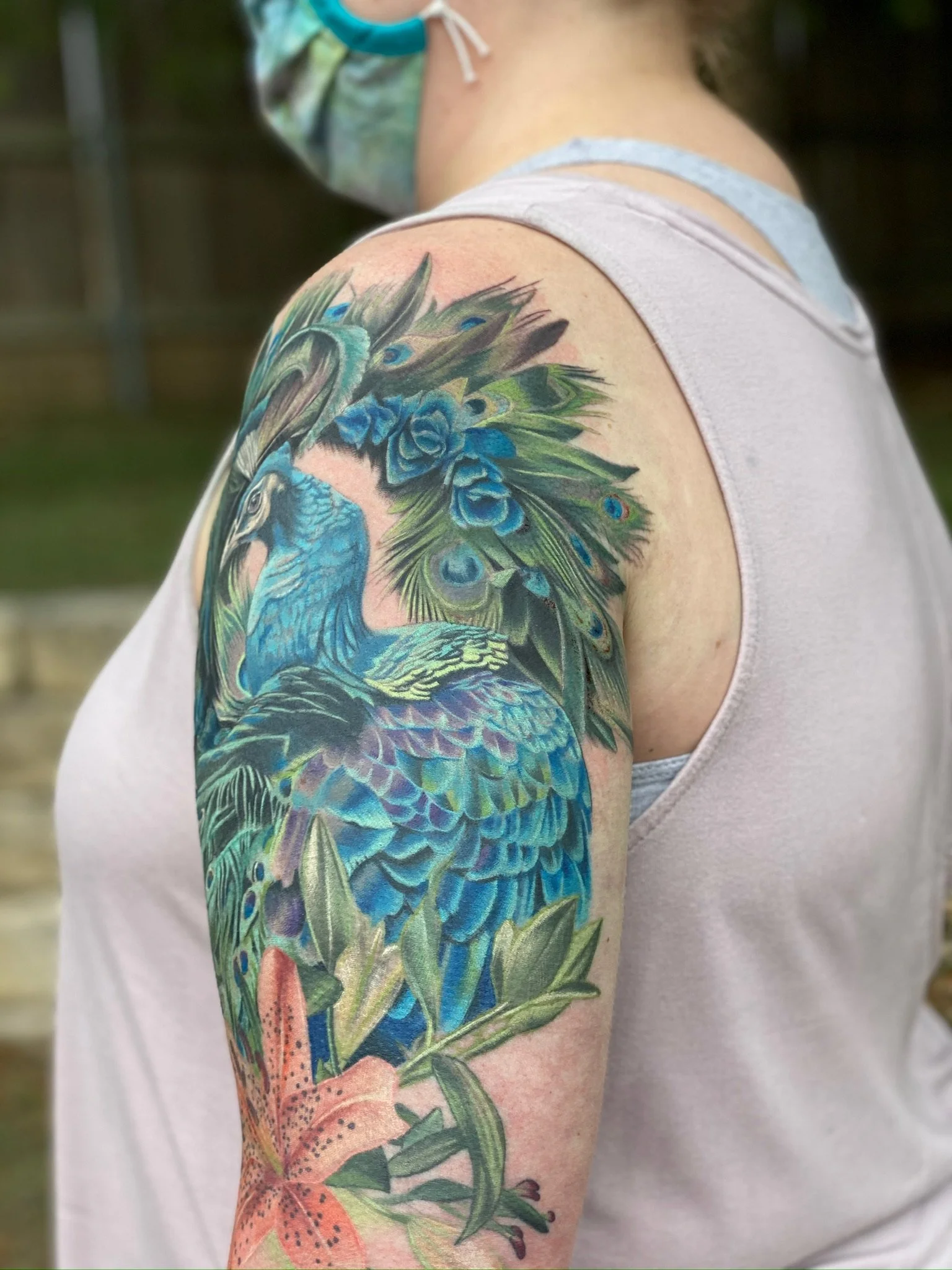 Tattoo of a blue peacock with detailed feathers, surrounded by green leaves and a pink lily on the upper arm of a person wearing a sleeveless shirt and a face mask.