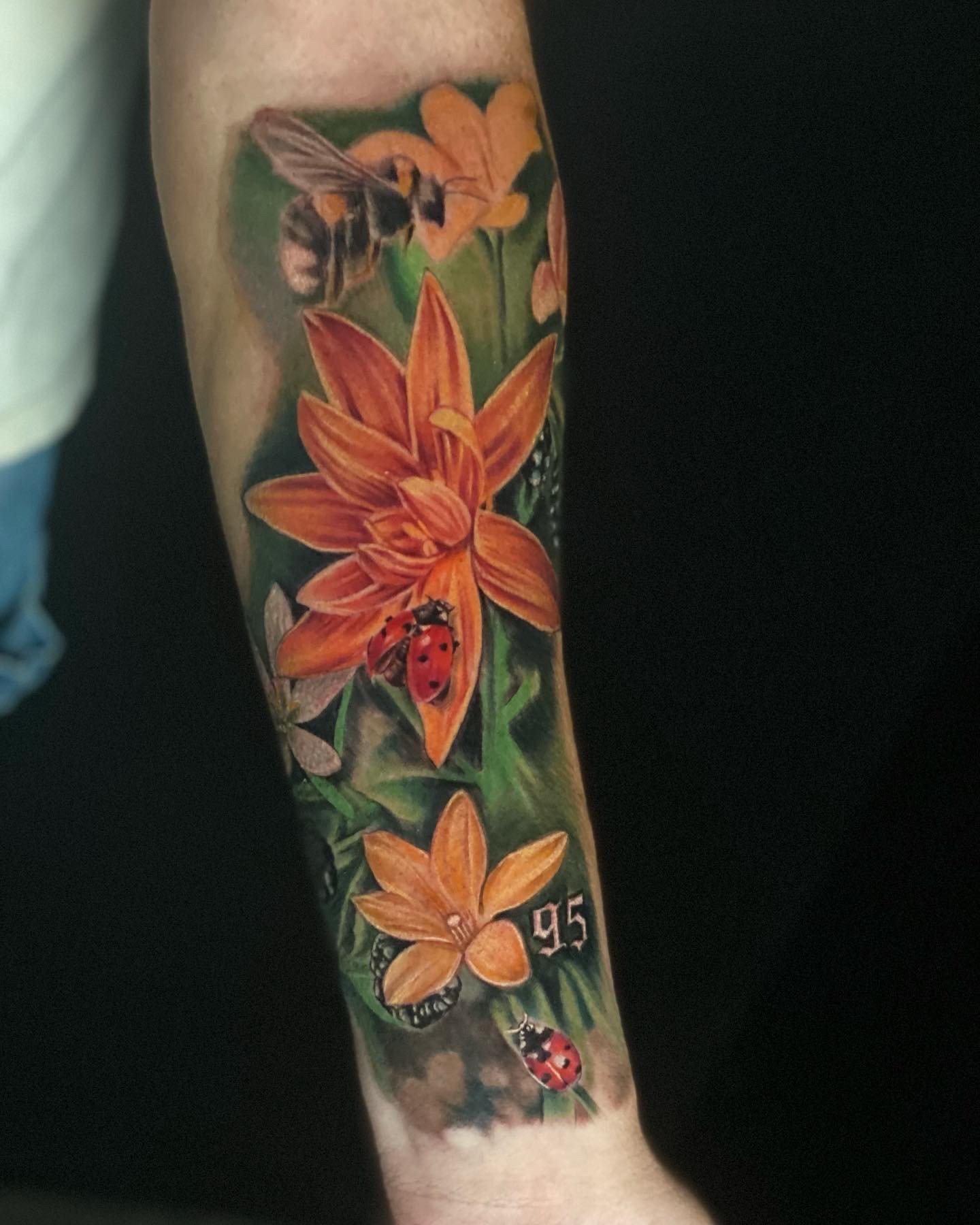 Tattoo of orange lilies flowers, a bee, and a ladybug on a person's arm.