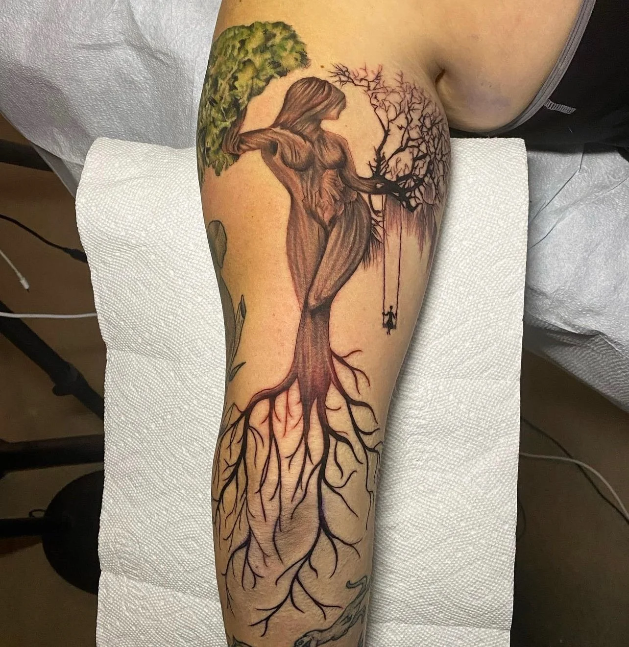 Tattoo of a tree with a woman’s figure forming the trunk, branches, and roots, and a swing hanging from one of the branches.