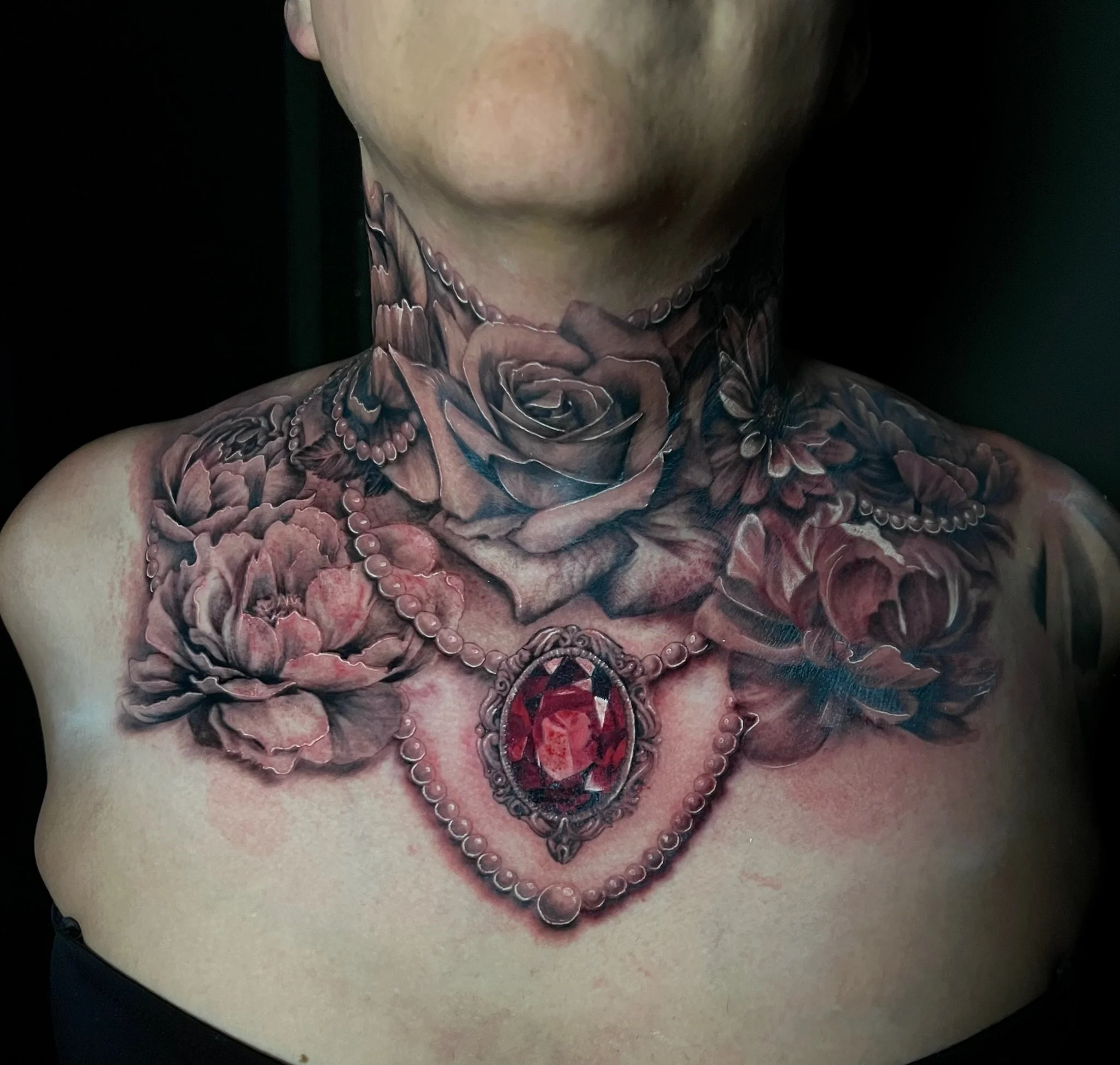 Tattoo of realistic roses, beads, and a large red gemstone pendant on a person's chest and neck.