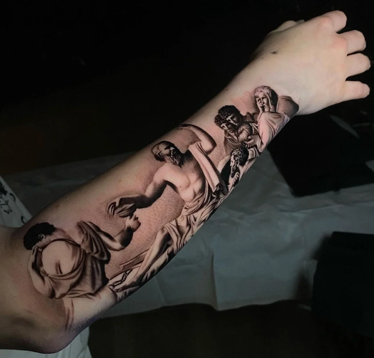 Black and gray realism tattoo of a classical scene on a person's forearm, depicting multiple figures in artistic poses and expressions.