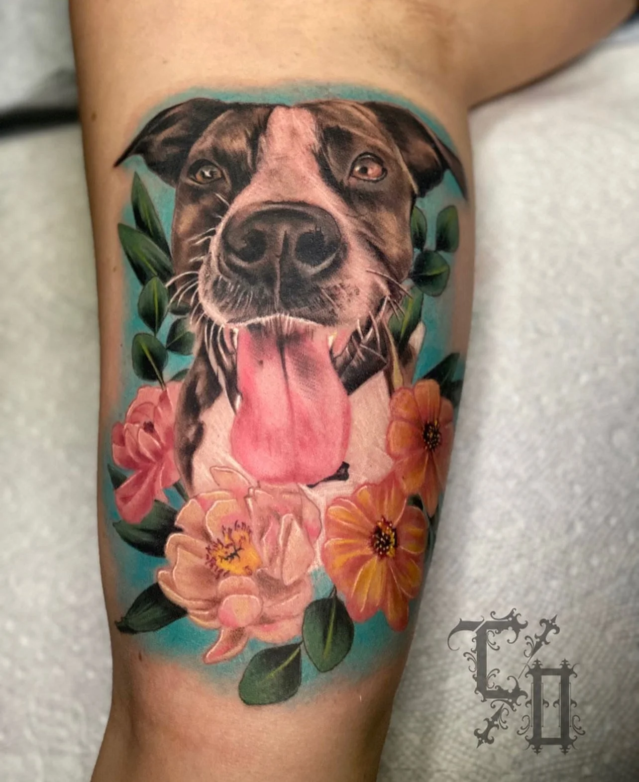 Realistic tattoo of a dog's face with its tongue out, surrounded by pink and orange flowers and green leaves.