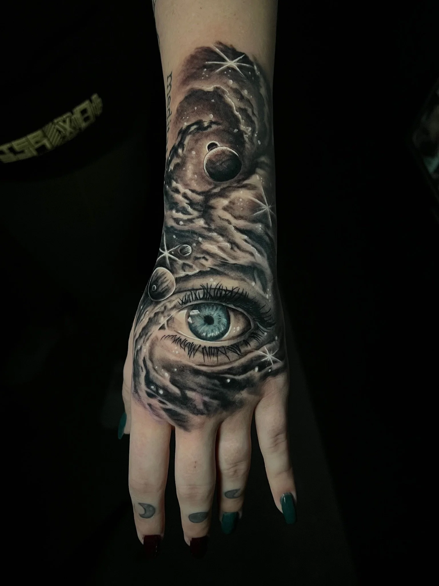 Tattoo of an eye surrounded by cosmic elements, including planets, stars, and swirling clouds, on a person's arm.