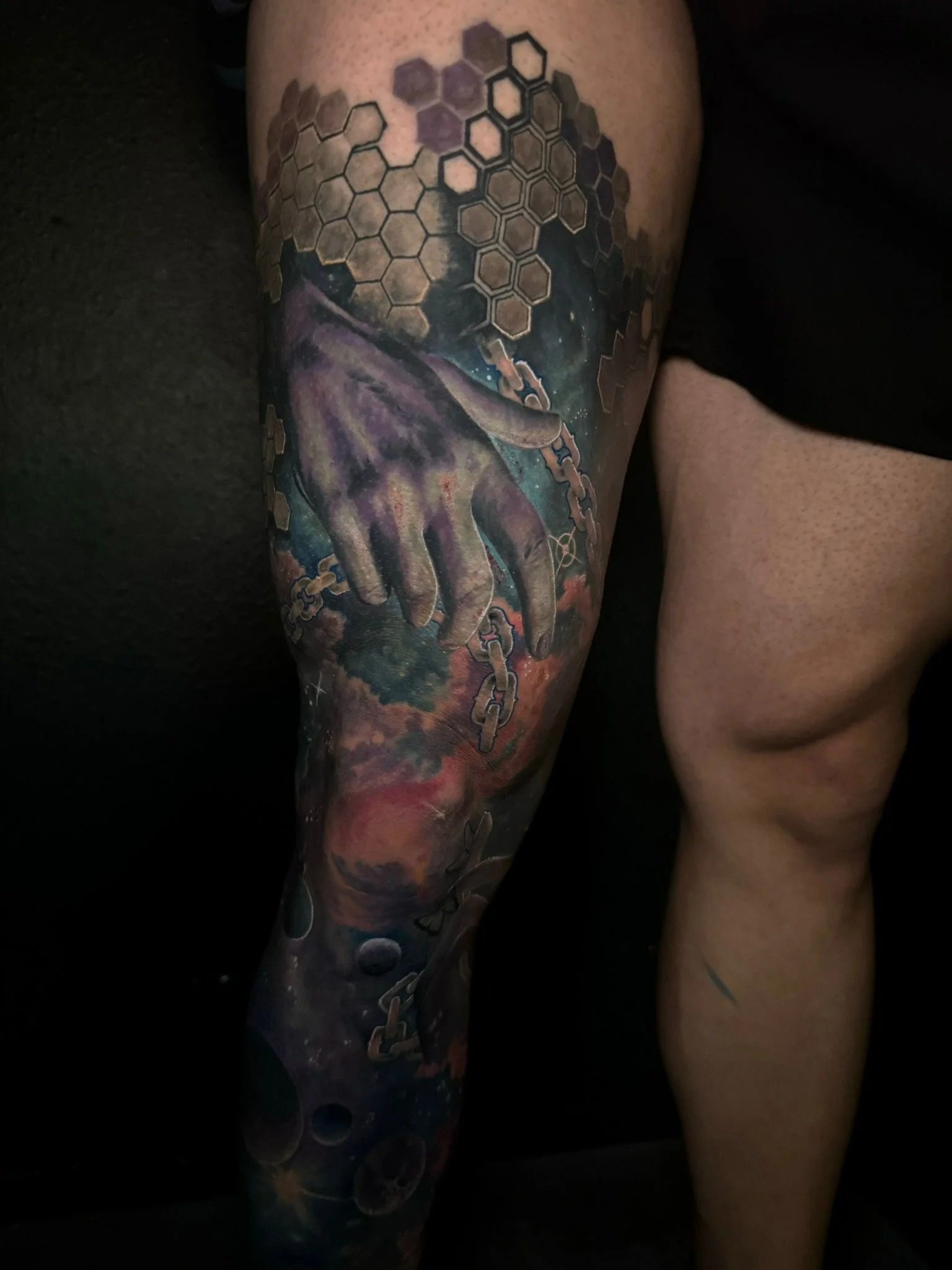 Tattoo of a cosmic scene with a purple hand reaching through space, surrounded by planets, clouds, and hexagonal patterns.