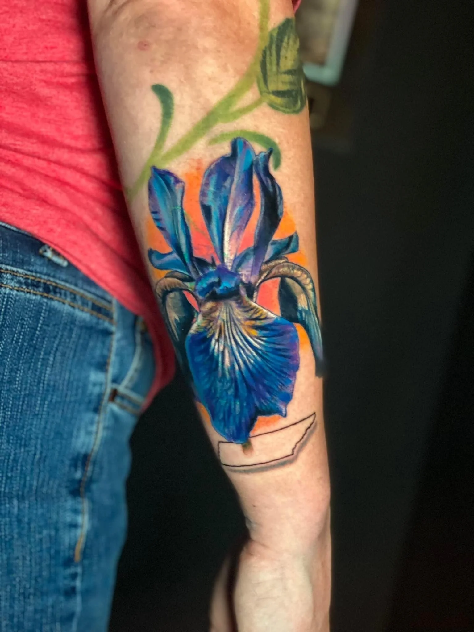 A colorful tattoo of an iris flower with green leaves on a person's arm, with an outline of the state of Arkansas below it.