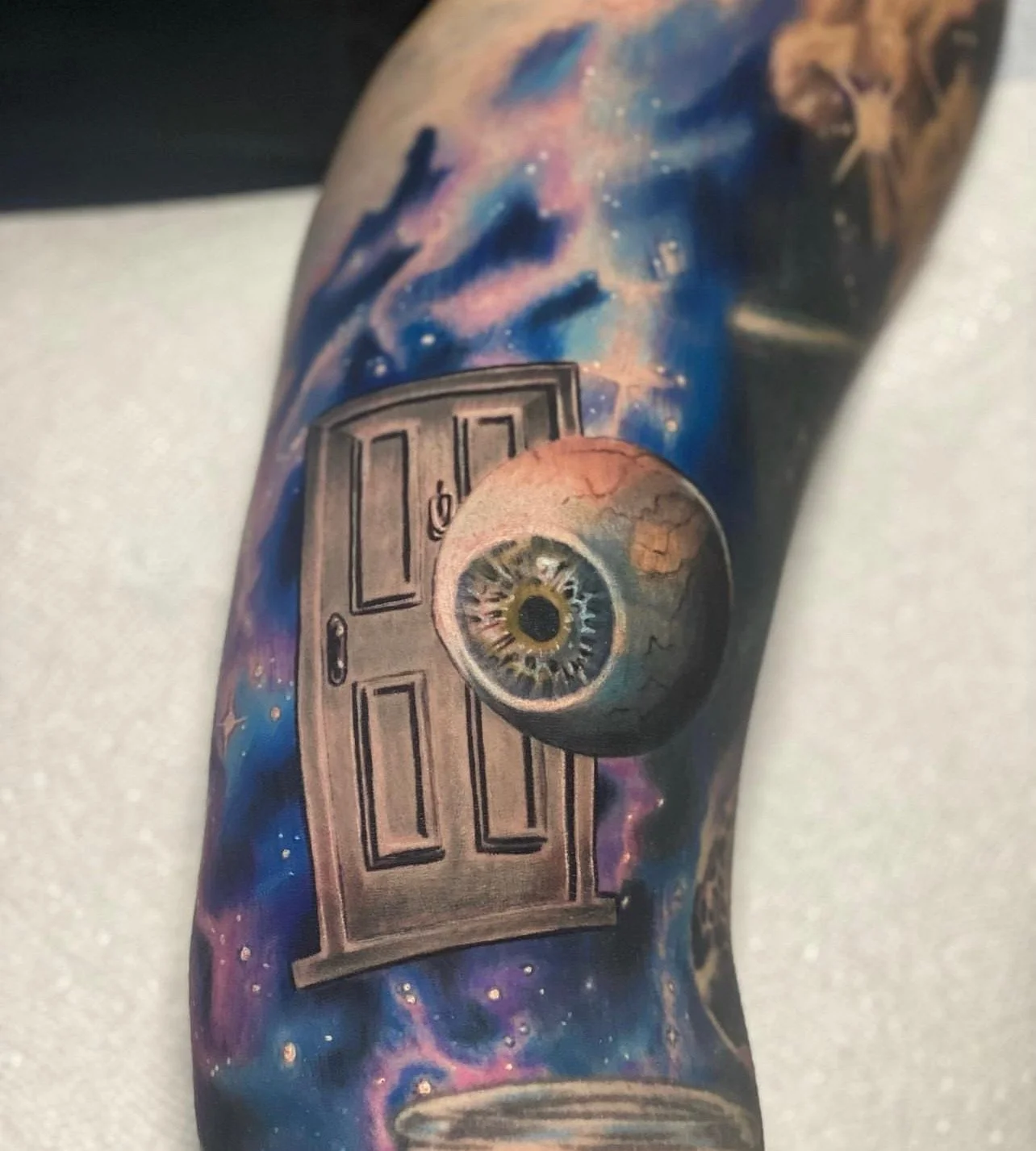 Tattoo of a door with a large eye in the center on a person's arm, with a cosmic background of stars and nebulae.