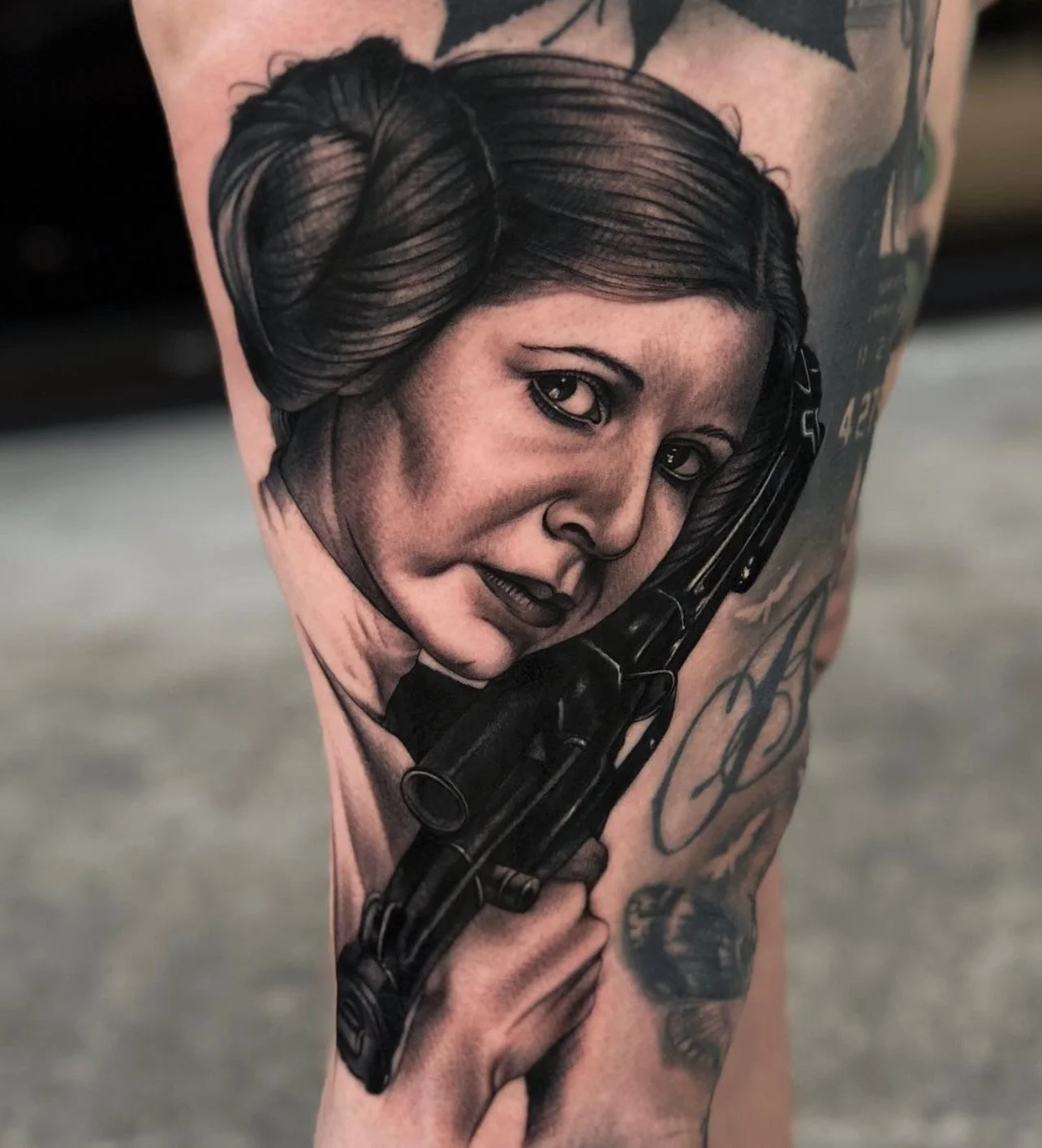 Black and gray tattoo of a woman's portrait with vintage hairstyle, holding a handgun, on a person's thigh.
