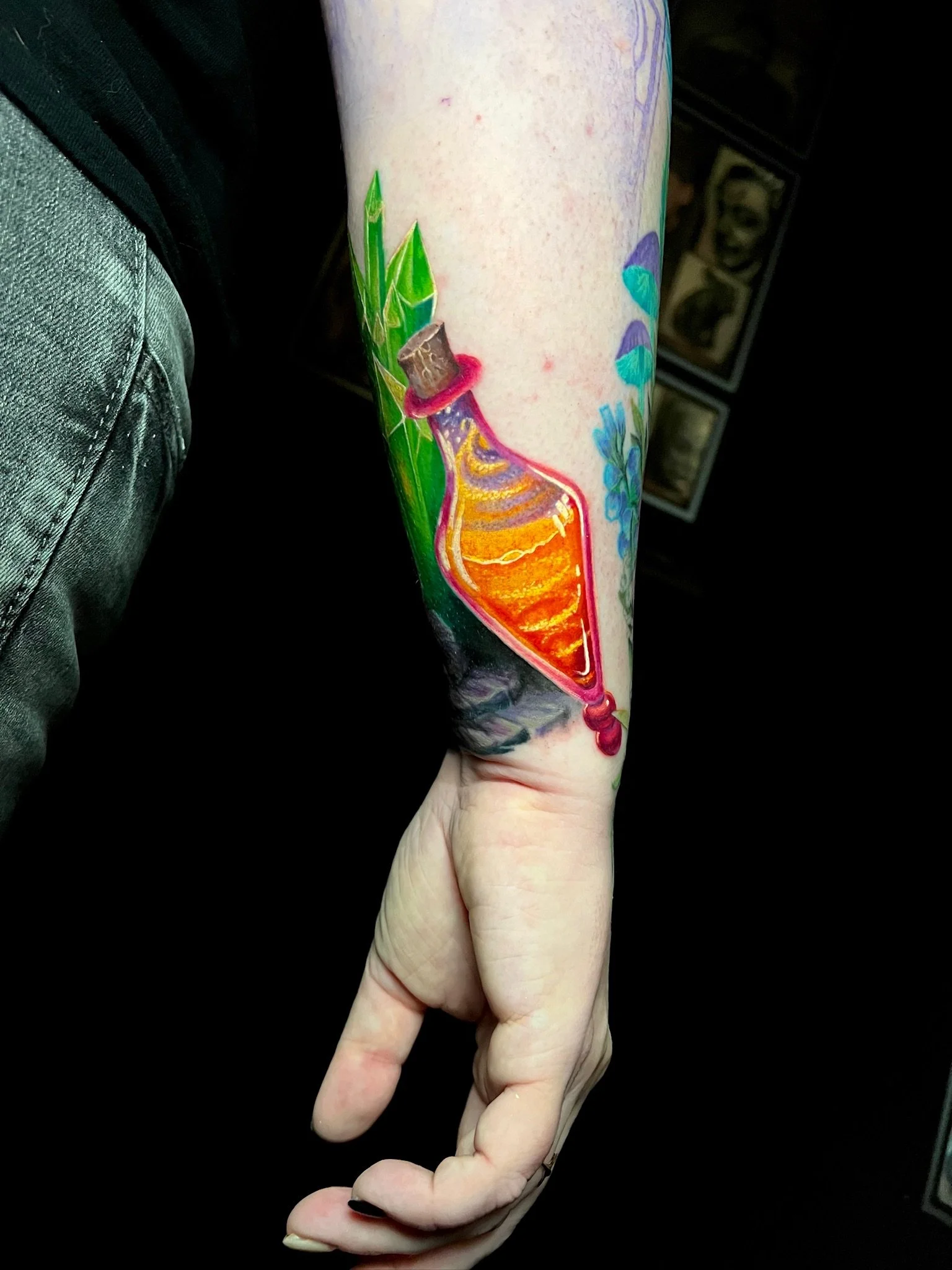 Colorful tattoo of a potion bottle with an orange liquid, green leaves, and a cork on the person's forearm.