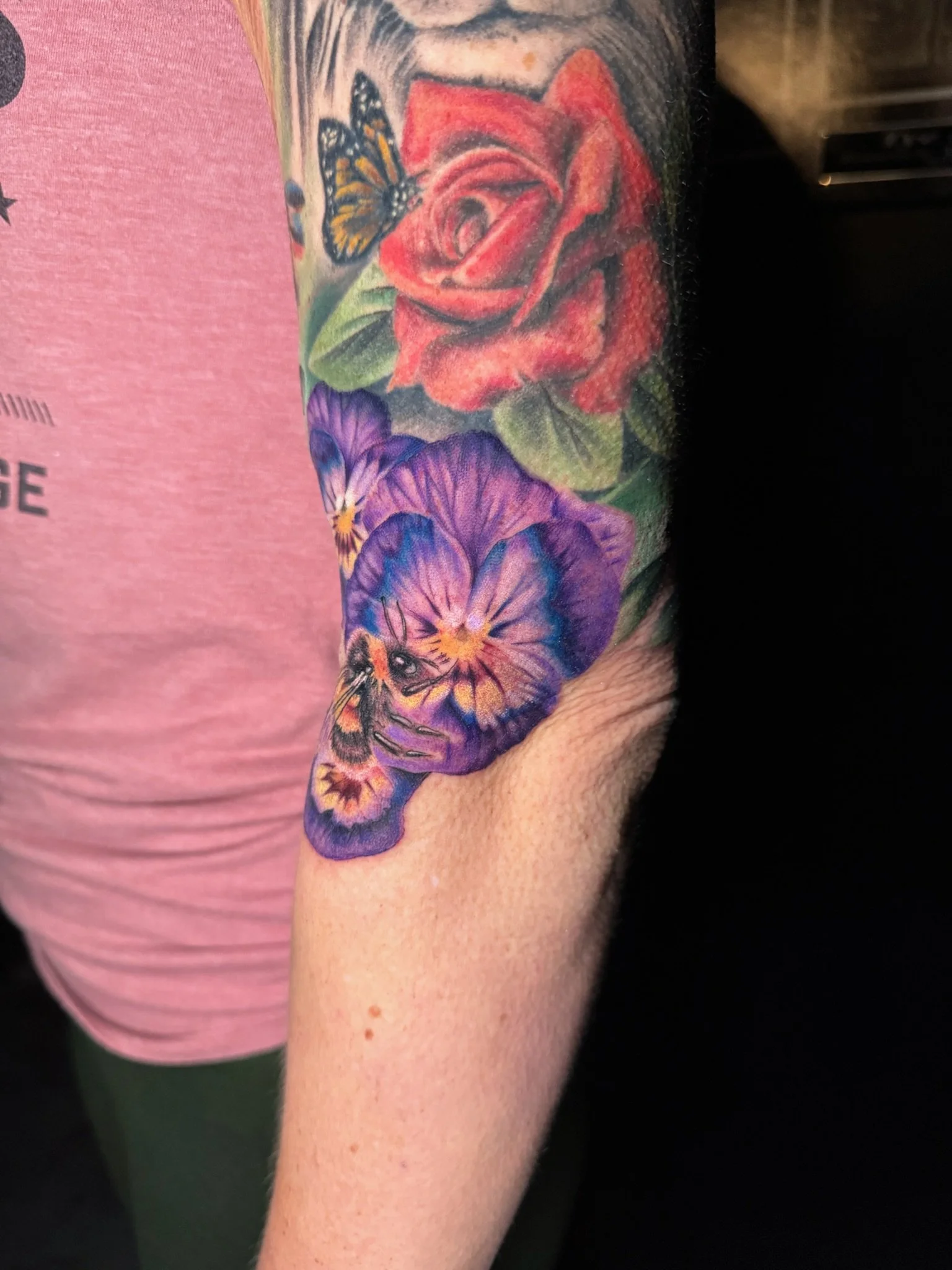Colorful tattoo on person's arm featuring a red rose, purple pansies, a butterfly, and a bee.
