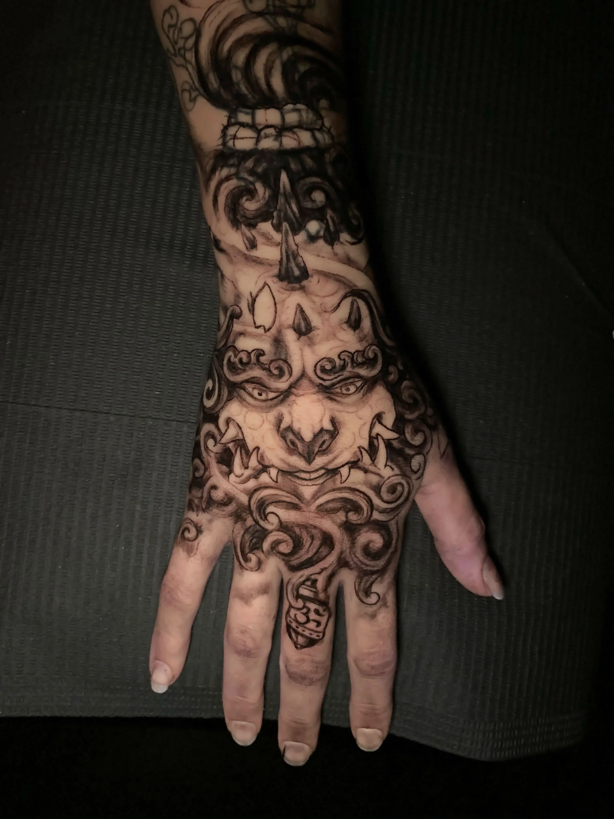 A person's hand with an intricate black tattoo design covering the wrist, hand, and fingers, featuring a skull, swirling patterns, and a large eye at the wrist, lying on a black textured surface.