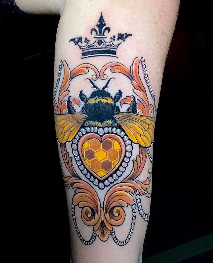 Colorful tattoo of a bee with honeycomb pattern inside a heart shape, topped with a crown, surrounded by decorative swirls and pearl-like beads.