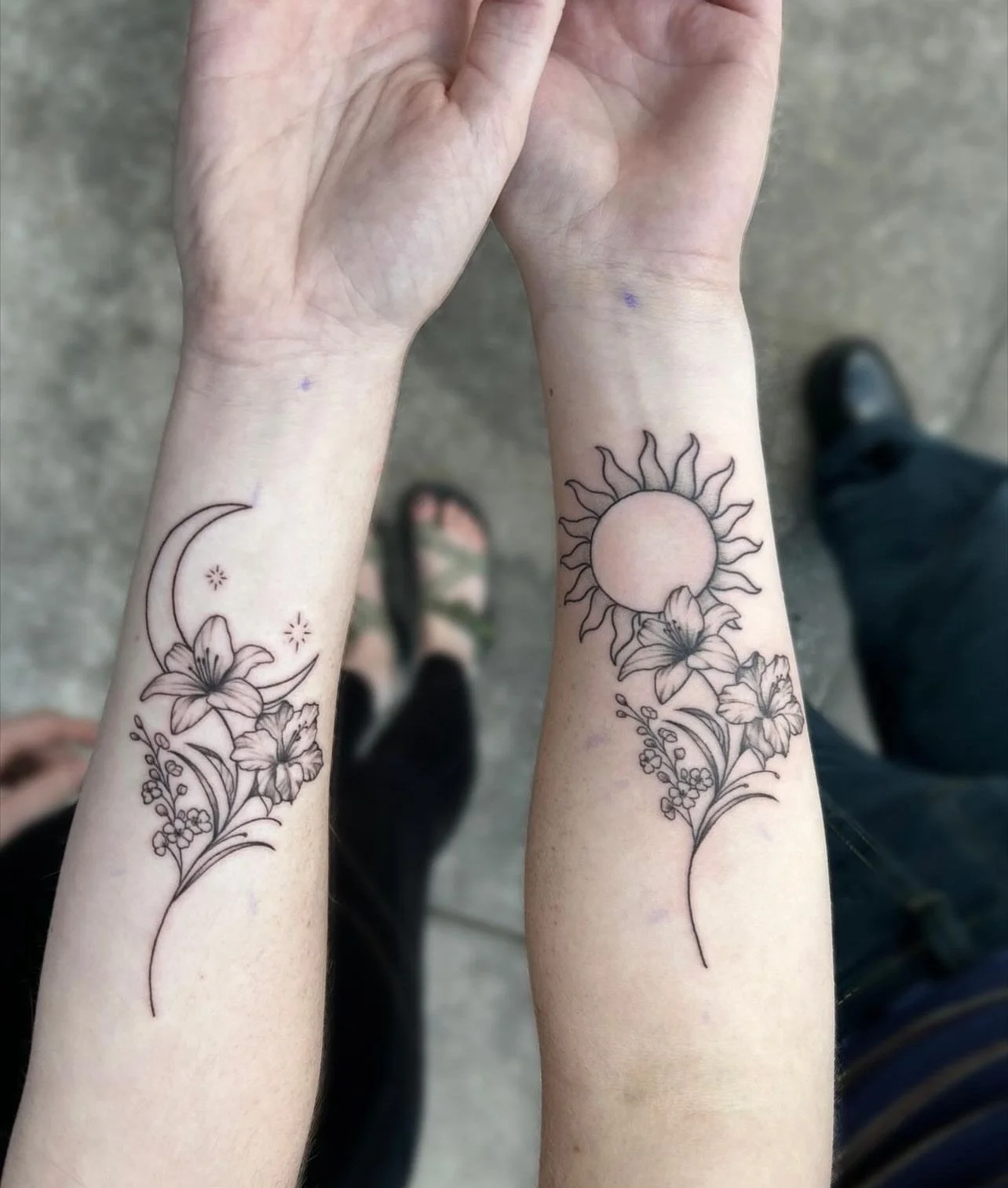 Close-up of two forearms with matching black line tattoos of flowers, a moon and starry sky, and a sun, with the person standing on a concrete surface.