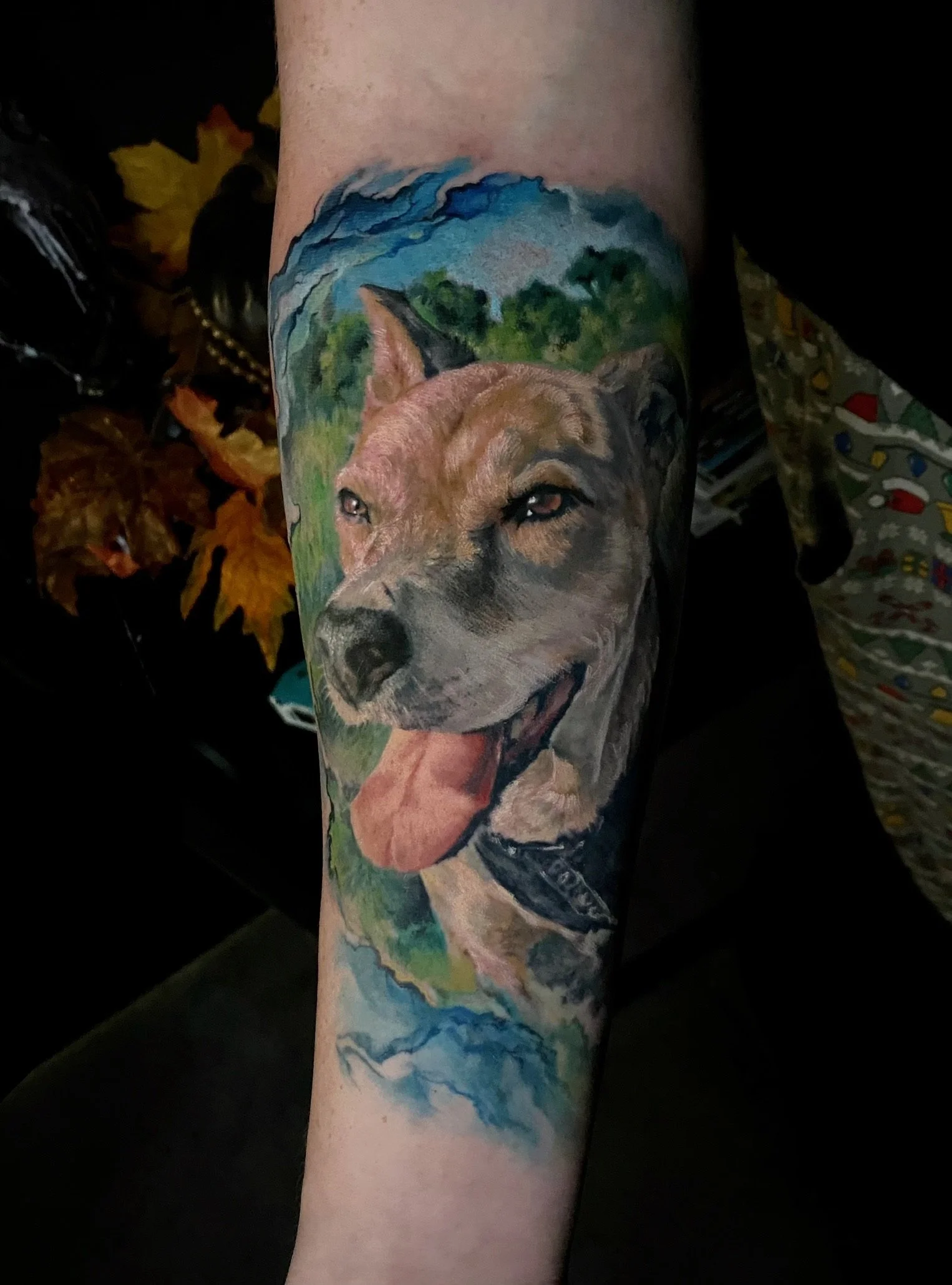Colorful tattoo of a happy dog with its tongue out, set against a landscape background with trees and mountains.