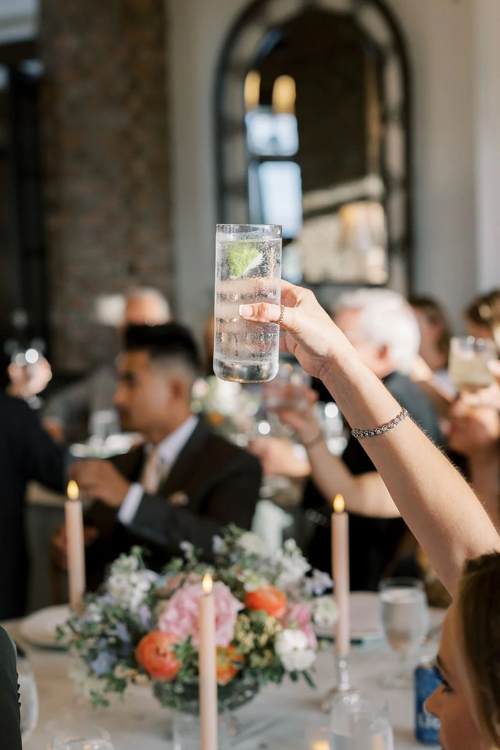 Person holding a glass of water with a lime wedge at a formal event, with blurred guests and floral centerpiece in the background.