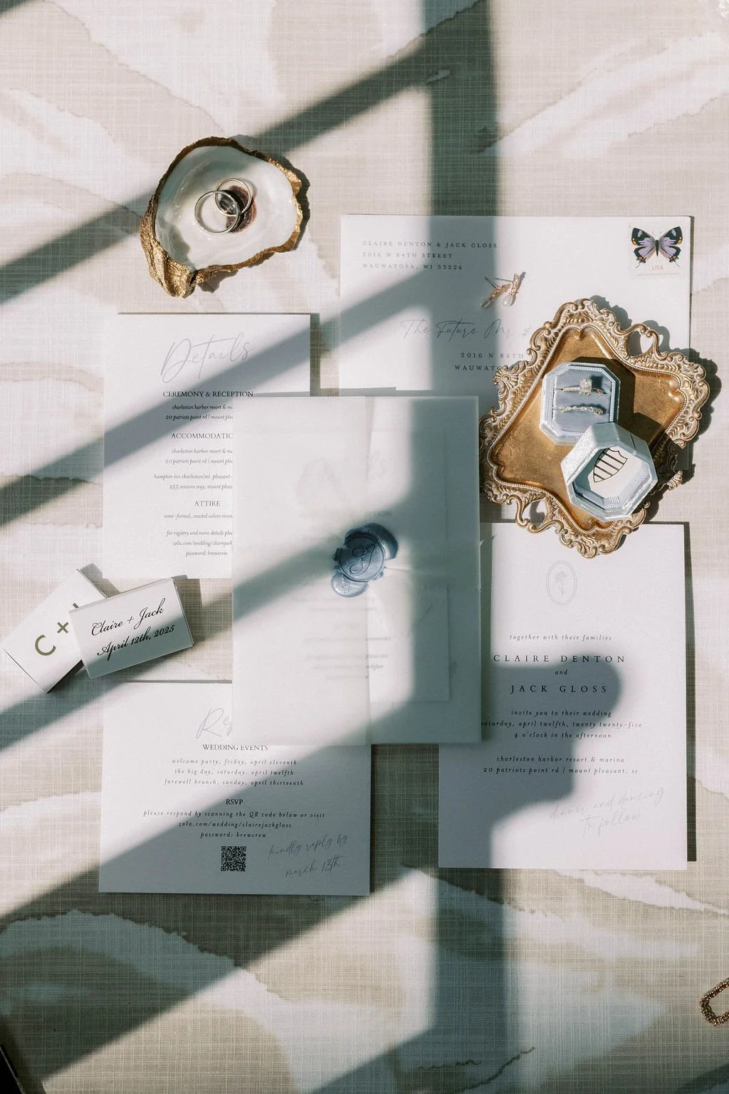 Wedding invitation suite and accessories arranged on a table, including stationery, rings, jewelry, and decorative items with shadows cast across the setup