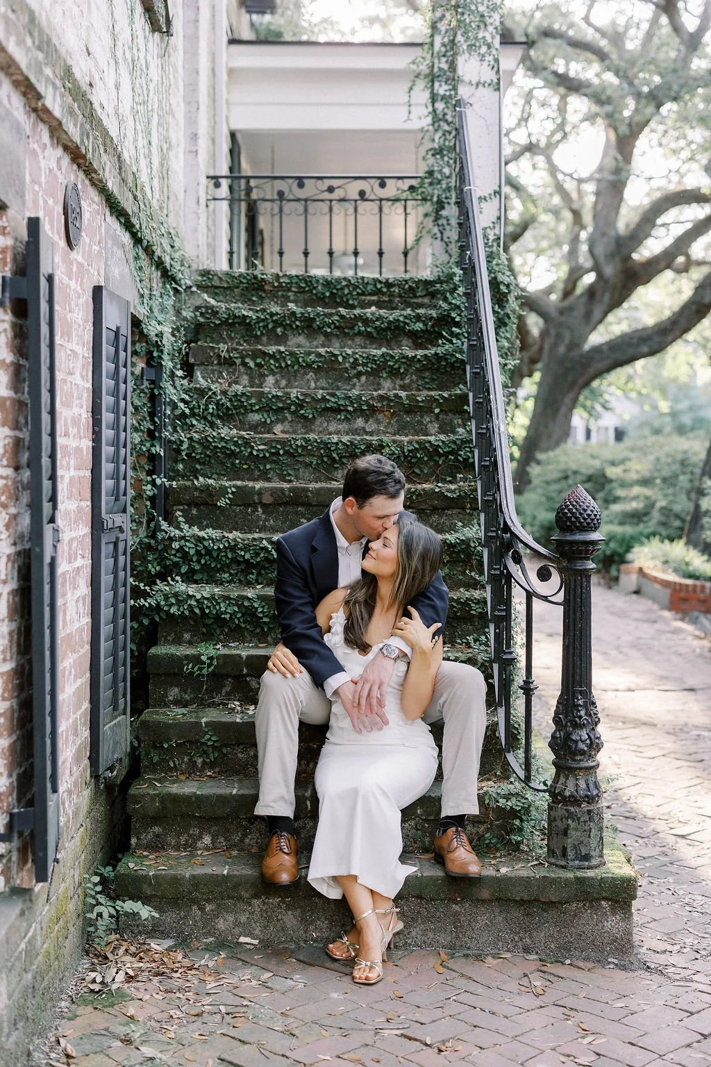 A couple sits on moss-covered steps, romantic moment with the man kissing the woman's forehead outdoors near trees and an old brick building.