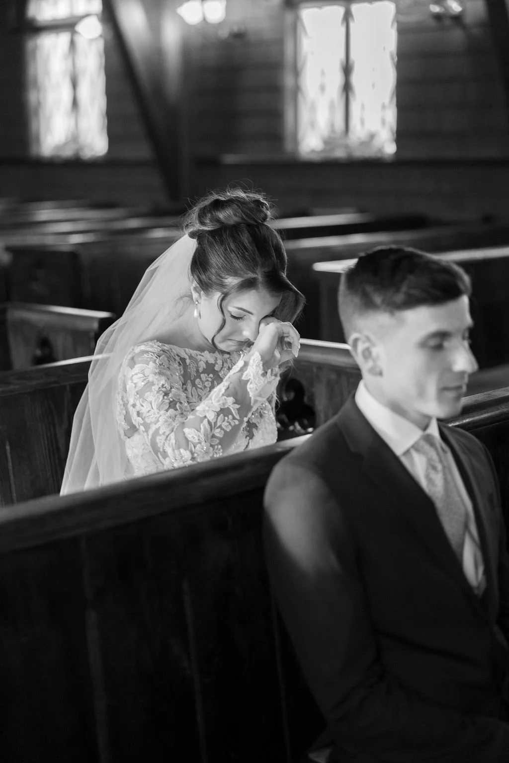 A bride dressed in a lace wedding gown and veil, sitting in a church, crying and wiping her tears while a groom in a suit sits in front of her, with a wooden pew separating them.