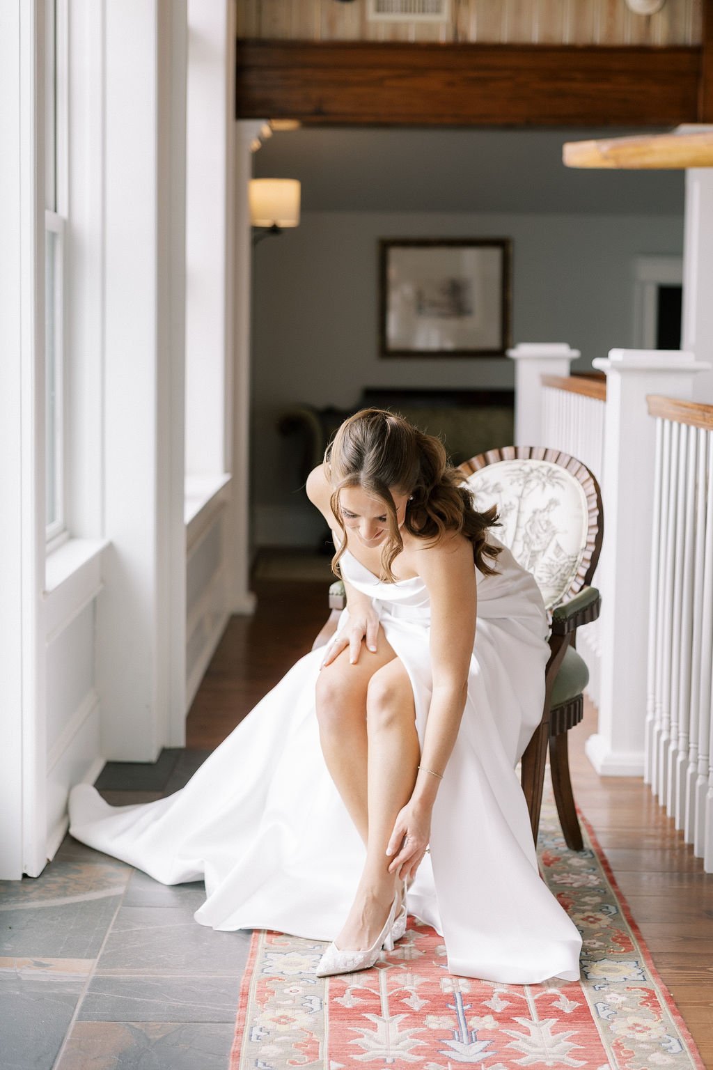 A bride sitting on a vintage wooden chair, putting on her high heel shoe, in a well-lit room with large windows and white trim.