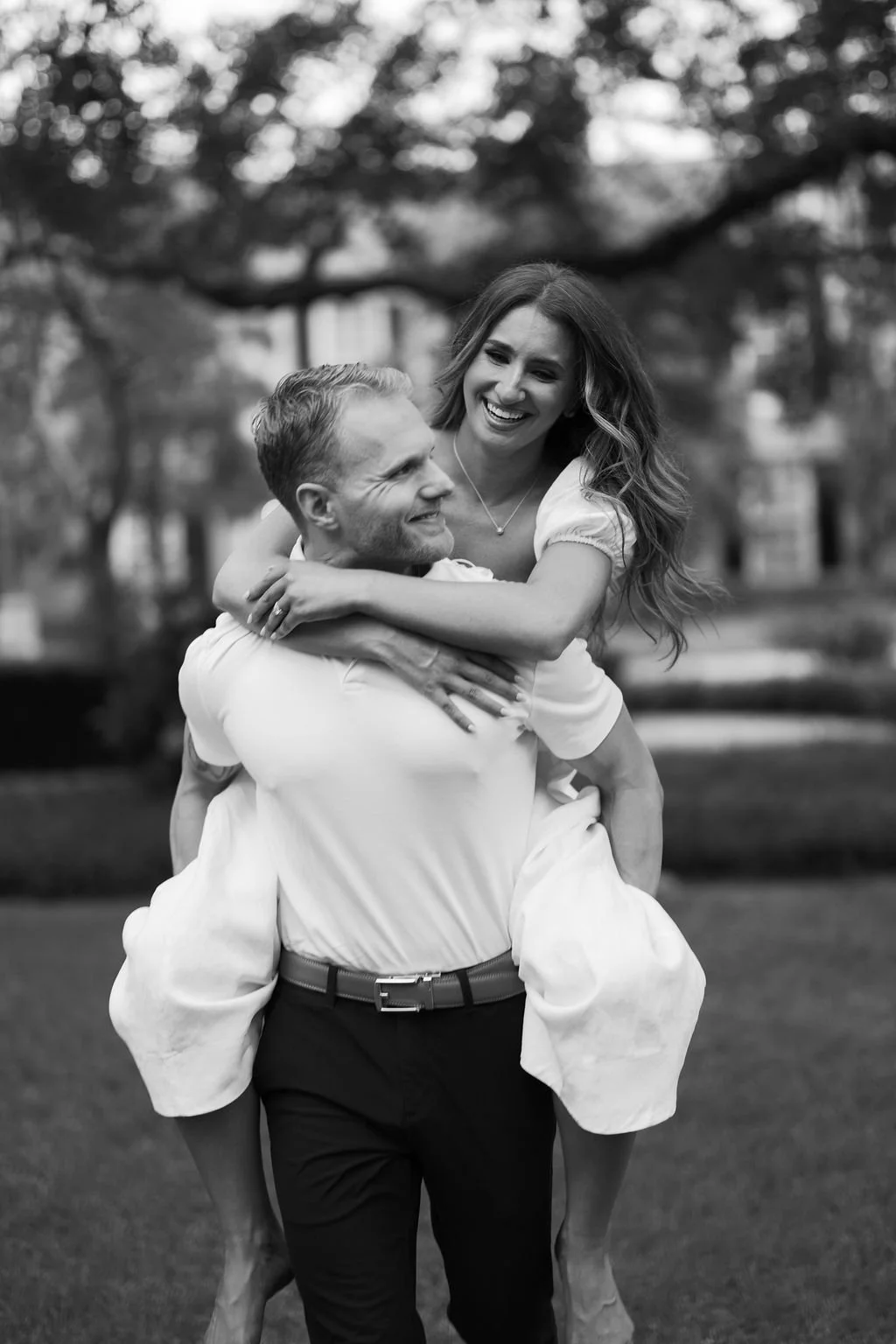 A happy couple outdoors in black and white, with the man carrying the woman on his back, both smiling and embracing in a park setting.