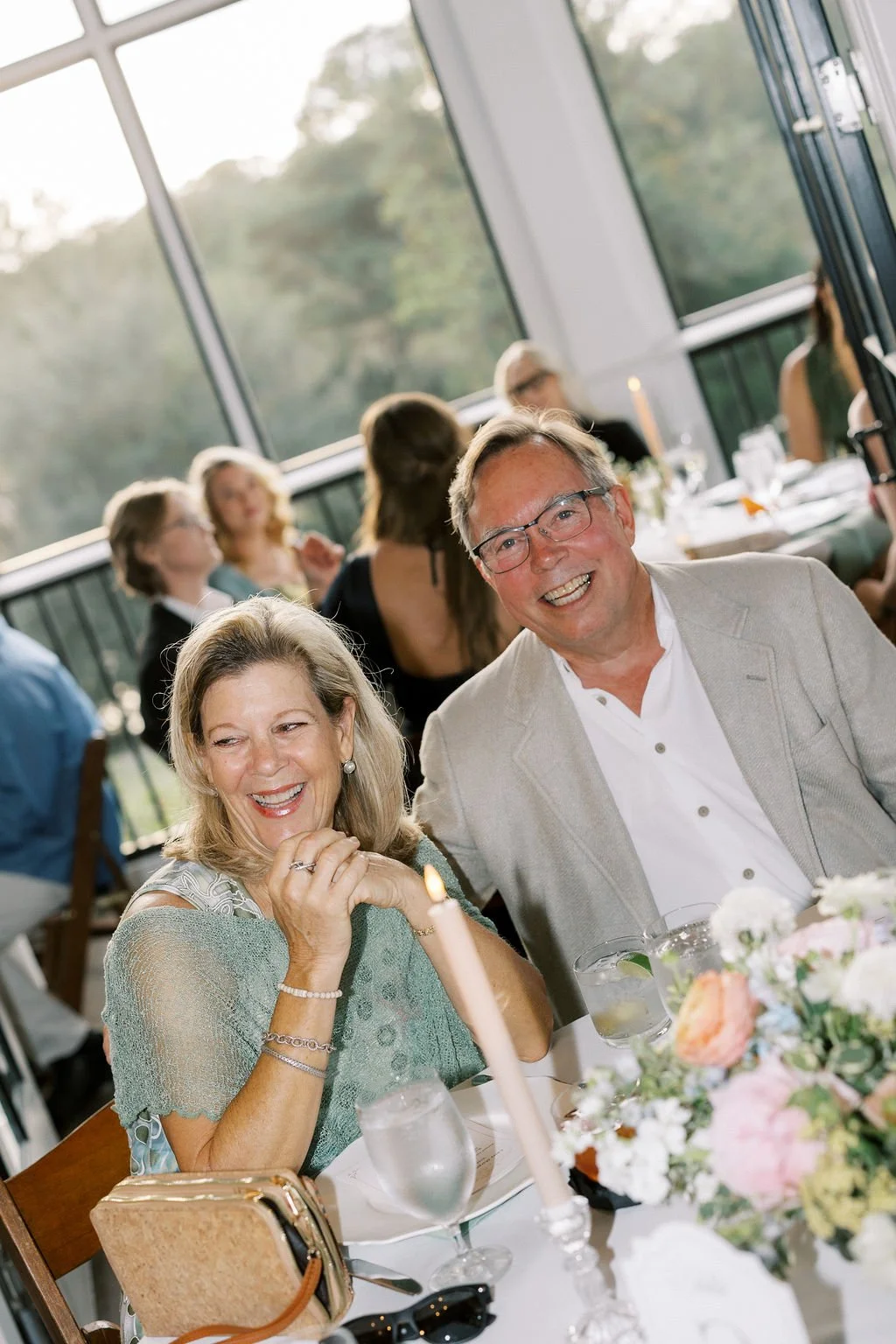 A smiling older couple, a woman and a man, sitting at a decorated table during a celebration in a bright room with large windows showing an outdoor view, surrounded by other guests and floral arrangements.
