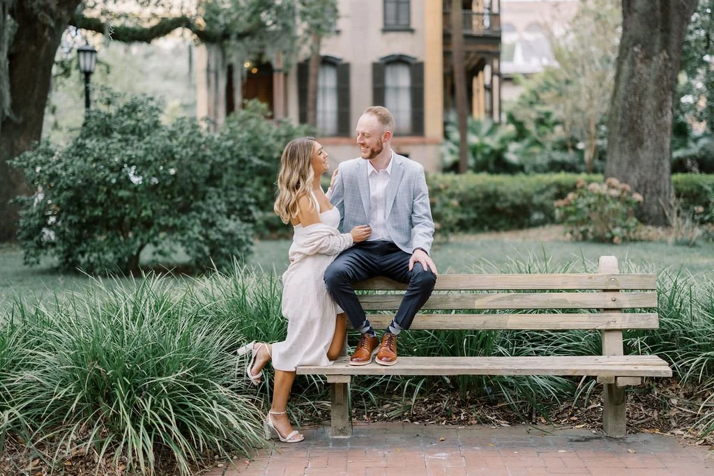 A woman and a man sit on a park bench, facing each other and smiling. The woman has long wavy hair and is wearing an off-shoulder white dress and heels. The man has a short beard, is wearing a light blazer, white shirt, dark pants, and brown shoes. T