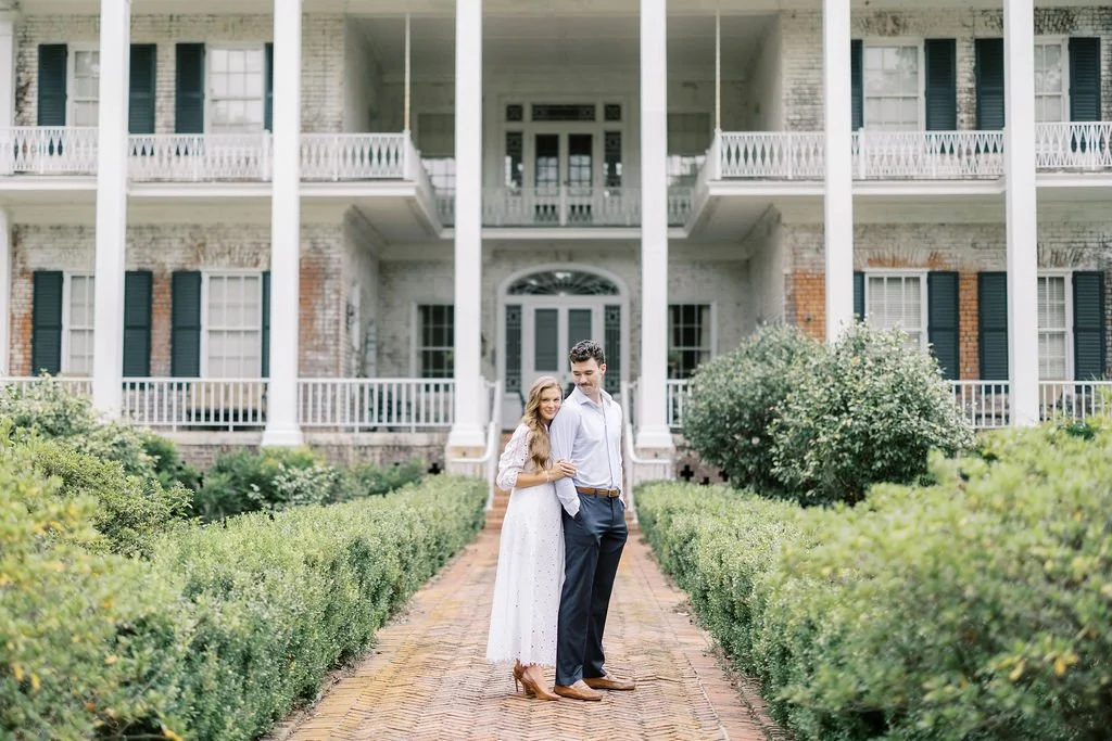 A couple standing close together on a brick pathway in front of a large, historic white house with columns and balconies, surrounded by well-maintained bushes and greenery.