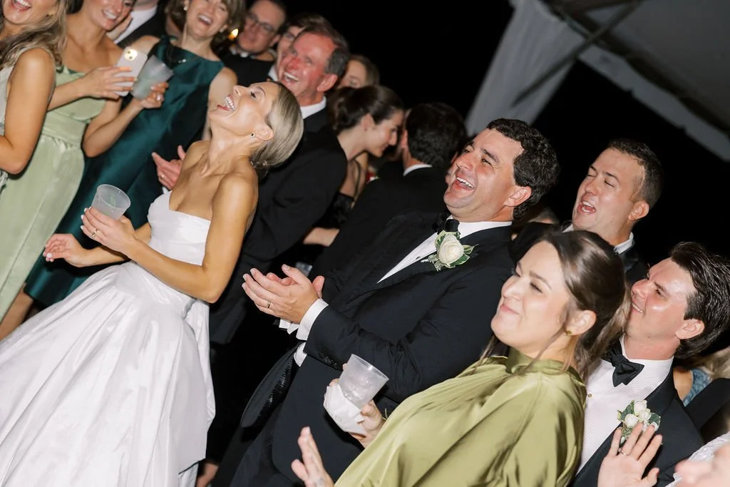 Group of people at a wedding reception, dressed in formal attire, smiling, laughing, and holding drinks.