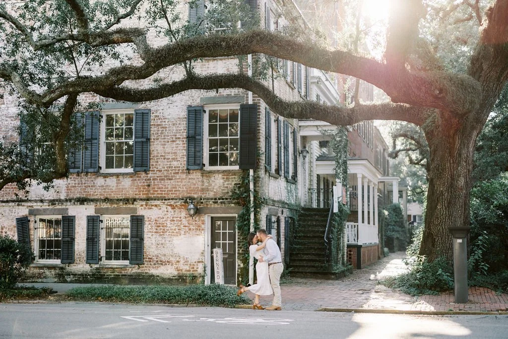 A couple kisses on a sidewalk in front of a historic brick building with blue shutters, a large tree overhead, and sunlight creating a warm atmosphere.