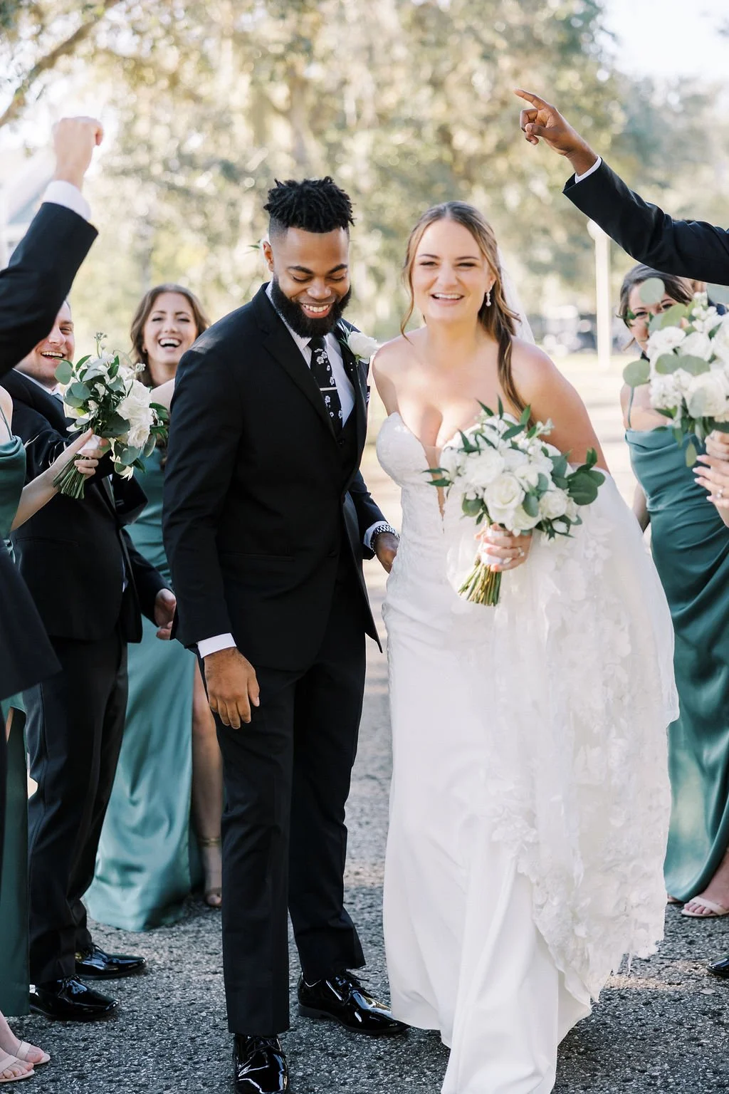 A wedding celebration with a bride and groom walking outdoors, surrounded by friends in formal attire, with the bride holding a bouquet of white flowers and smiling.