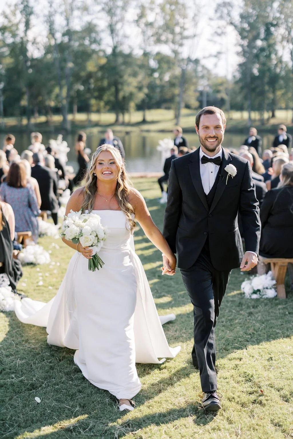 A newlywed couple walking hand-in-hand outdoors at their wedding ceremony, with guests seated and trees in the background.