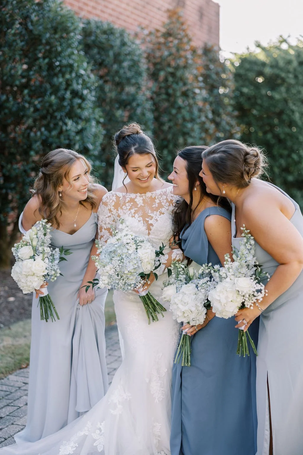 Group of five women, including a bride in a wedding dress and four bridesmaids in blue dresses, smiling and holding bouquets of white flowers outdoors.