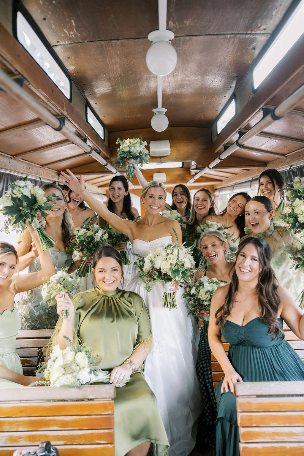 A group of women, including a bride in a white wedding dress, celebrating on a bus with bouquets of flowers, smiling and waving.